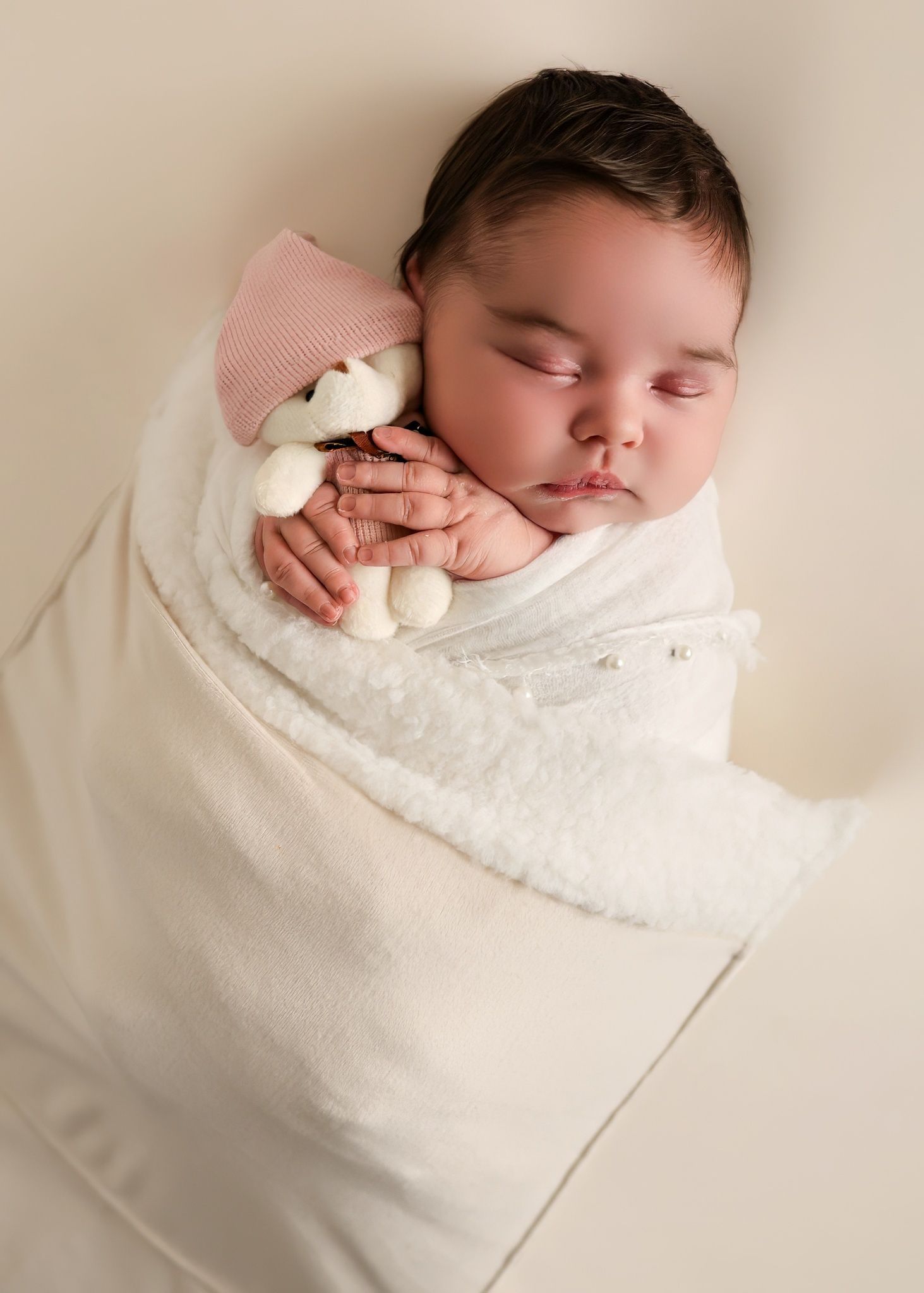 Sleeping newborn wrapped in white blanket with small plush toy on white background.