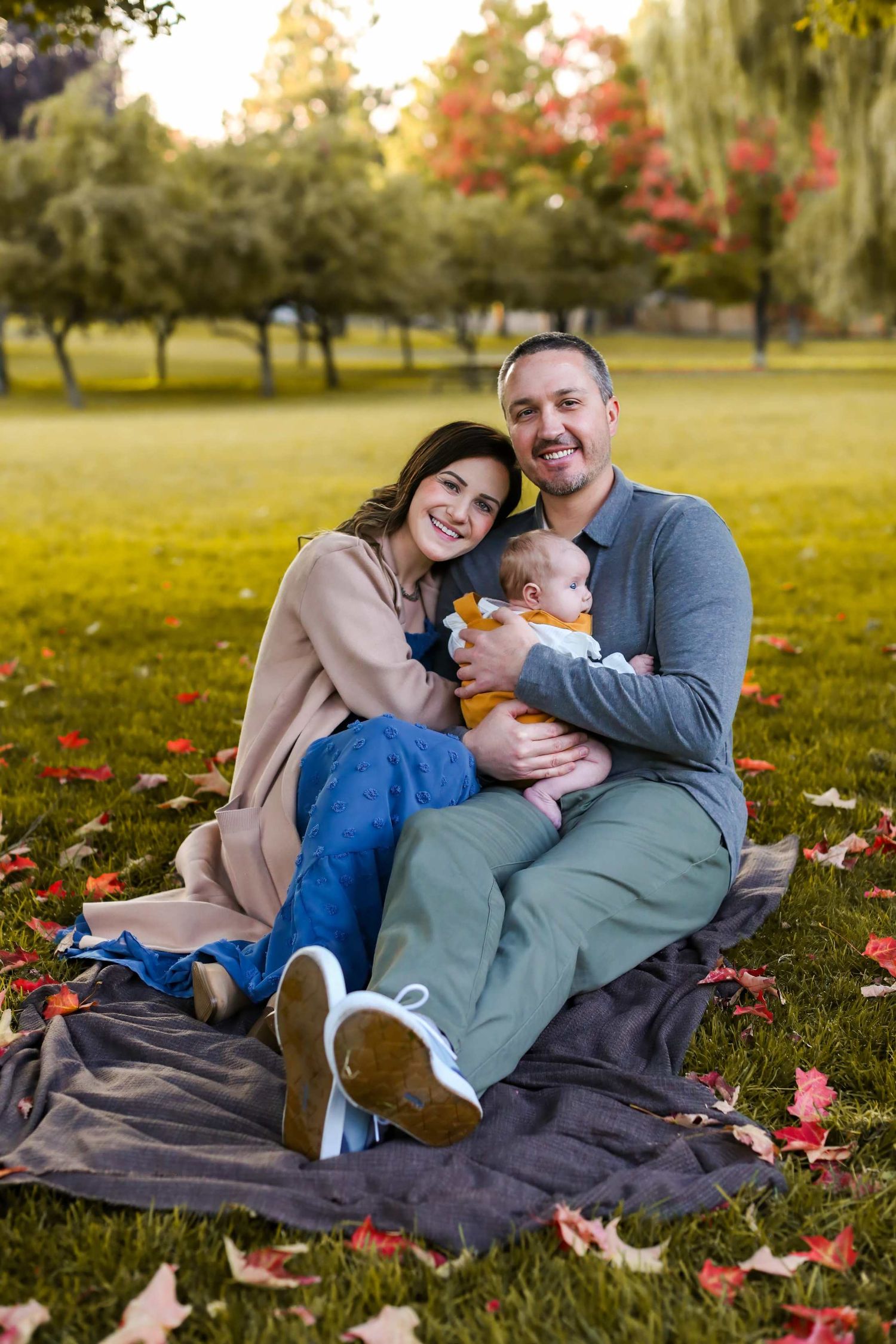 A smiling couple sits on a blanket in a park holding their newborn baby surrounded by colorful autumn leaves.