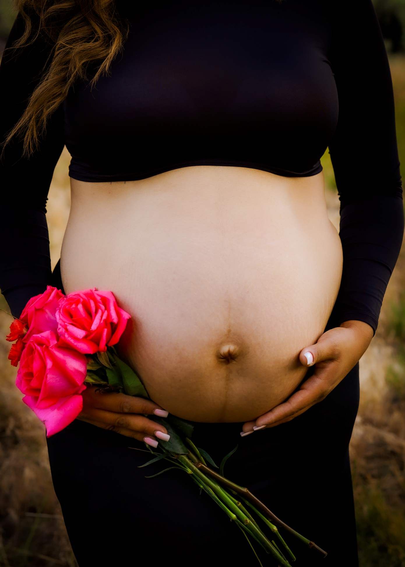 A pregnant belly in black clothing holds bright pink roses against a dark background. Indian Ford Meadows, Sisters Oregon