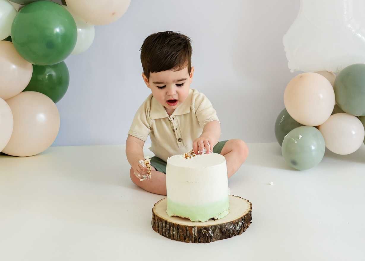 A baby in beige clothing reaches toward a mint green cake with white decorations and balloon backdrop.