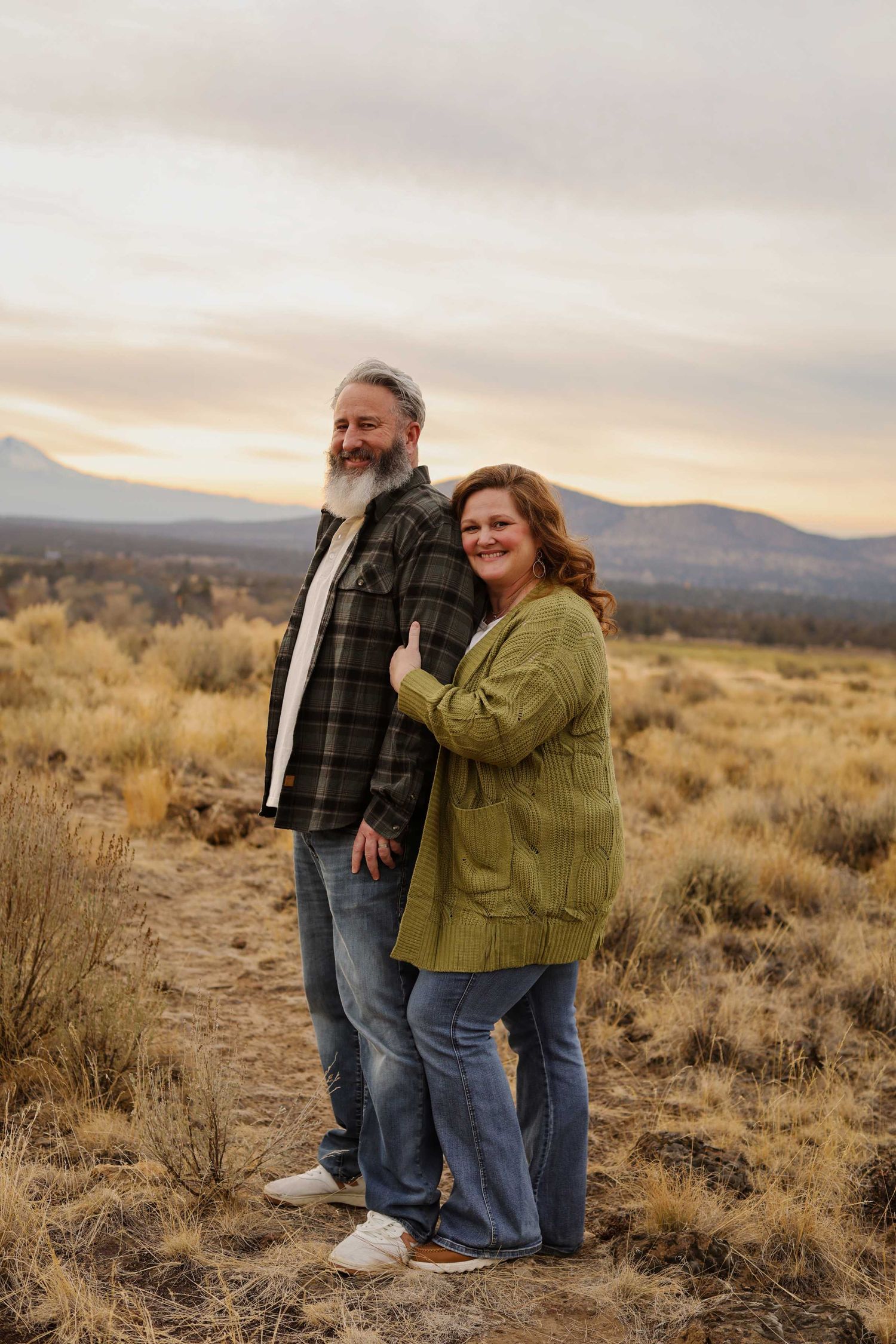 A couple poses together in a desert landscape with mountains visible in the background at sunset.