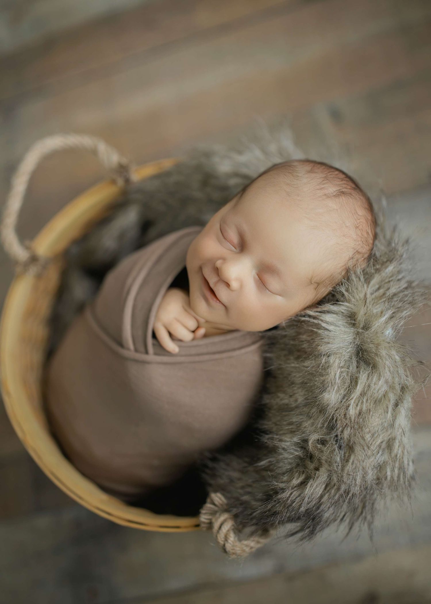 Sleeping newborn wrapped in brown swaddle nestled in furry basket with wooden handles.