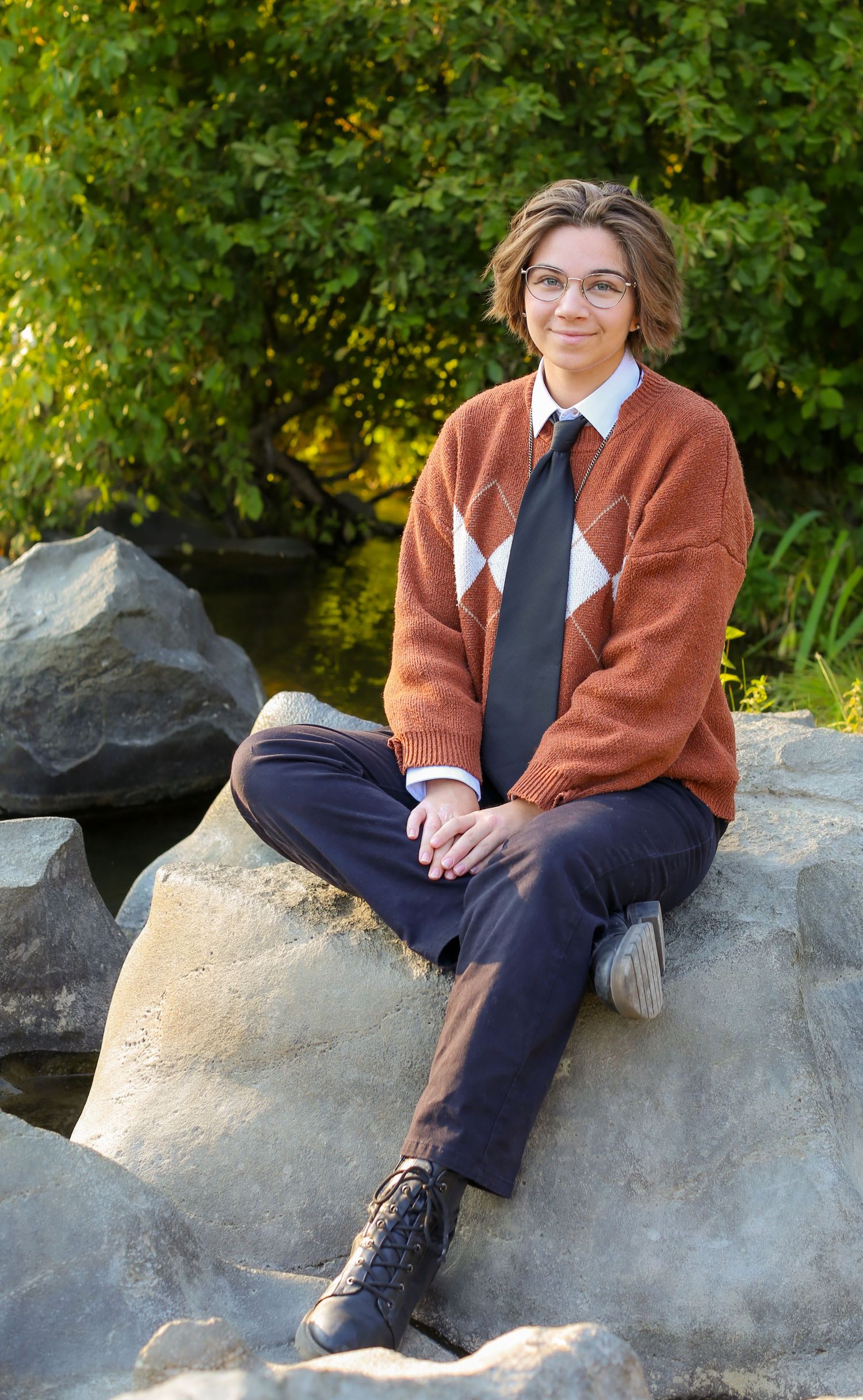 Person in rust argyle sweater and tie sits on rocks outdoors, smiling, with lush green trees in background.