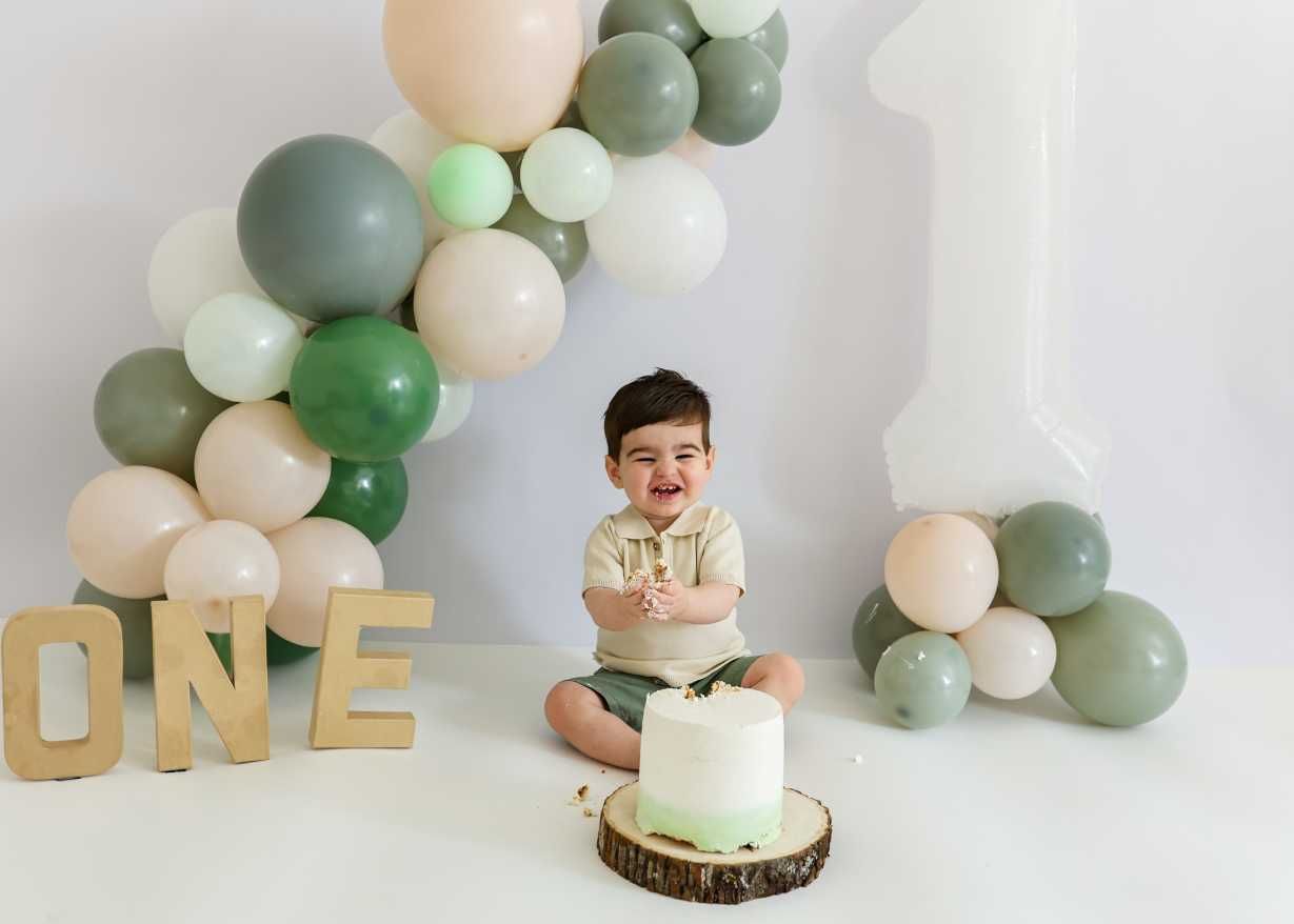 Smiling baby boy celebrates first birthday with green and beige balloon arch, ONE letters, and white smash cake.