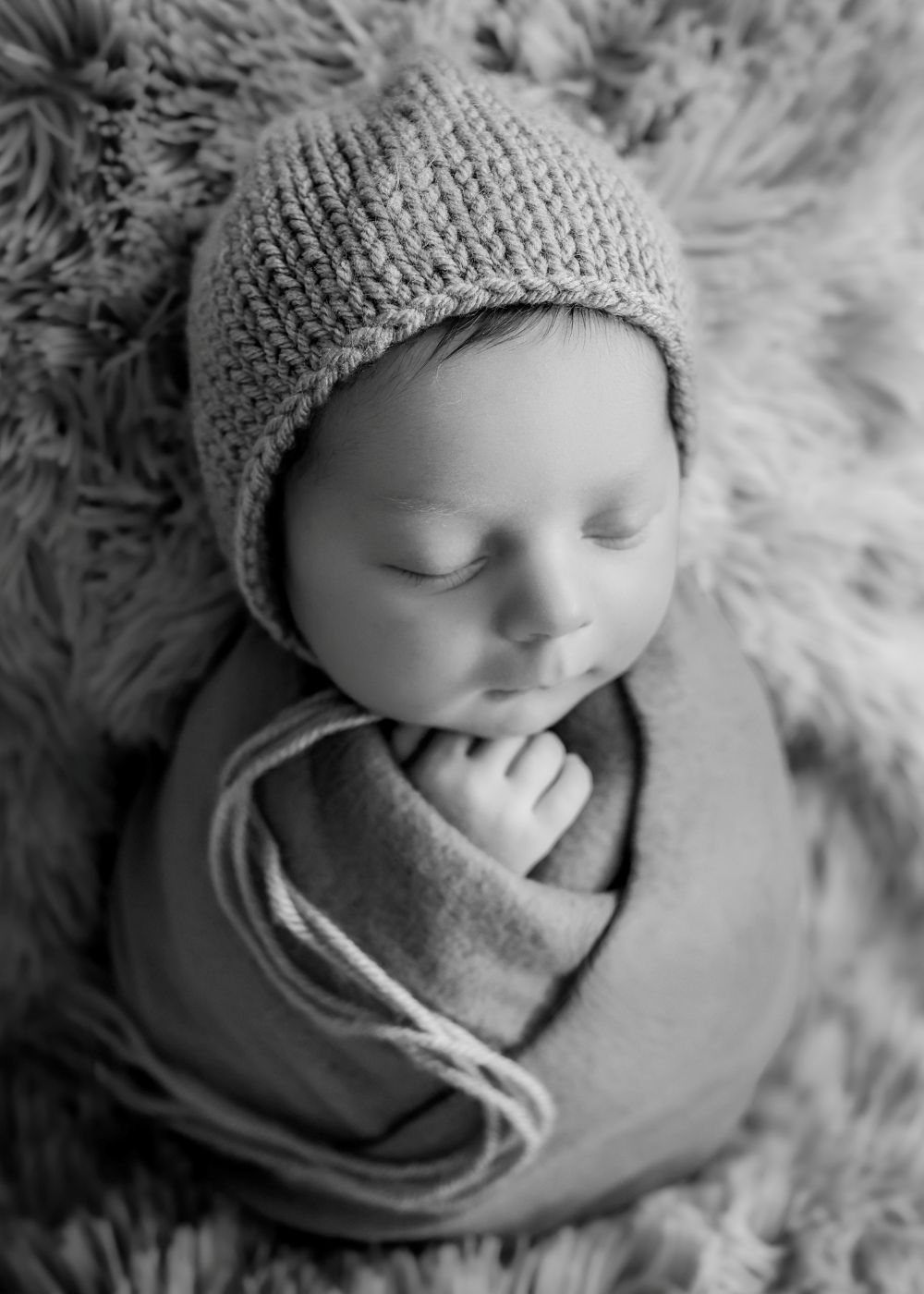 Black and white newborn photo of sleeping baby swaddled in soft wrap wearing knit bonnet on fluffy rug.