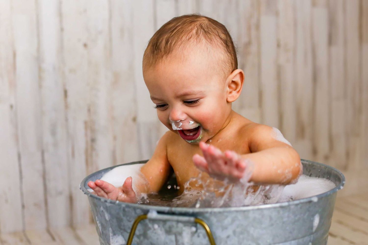Happy baby splashing and laughing during bath time in metal wash basin against rustic wooden wall.