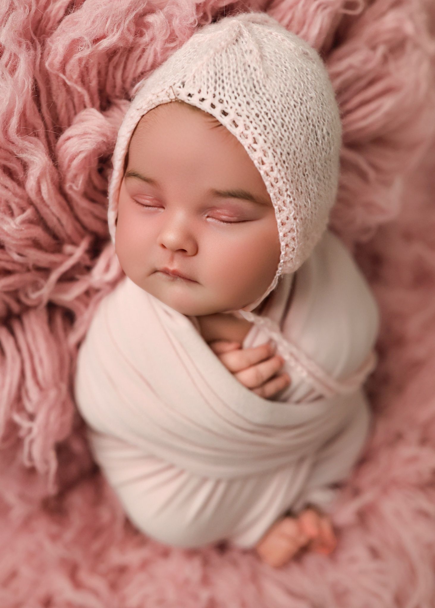 Peaceful sleeping newborn swaddled in white fabric wearing a delicate knit bonnet against soft pink textured backdrop. Redmond Oregon Studio