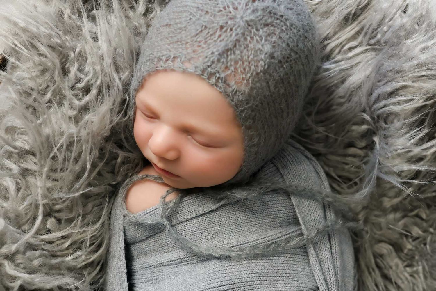 Baby resting on plush gray fur blanket wearing knitted bonnet in monochromatic portrait.