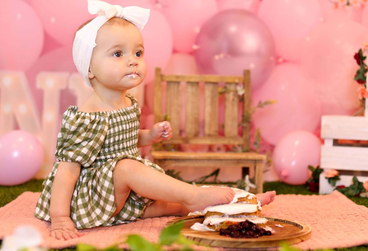 Adorable baby girl in gingham dress sits beside smash cake with pink balloon backdrop.