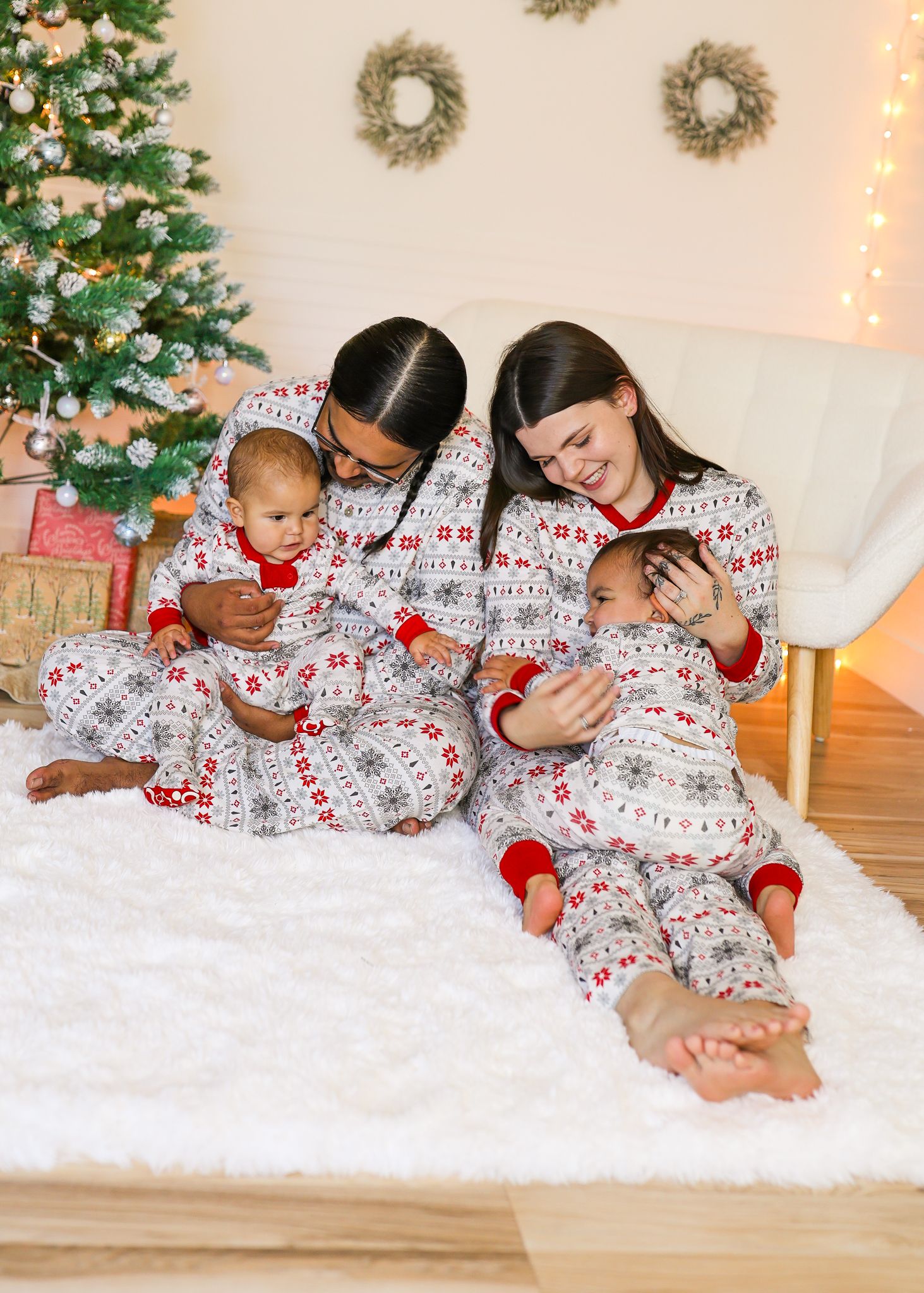 Two women and two babies in matching red and white Fair Isle holiday pajamas sit on a white faux fur rug beside a decorated Christmas tree.
