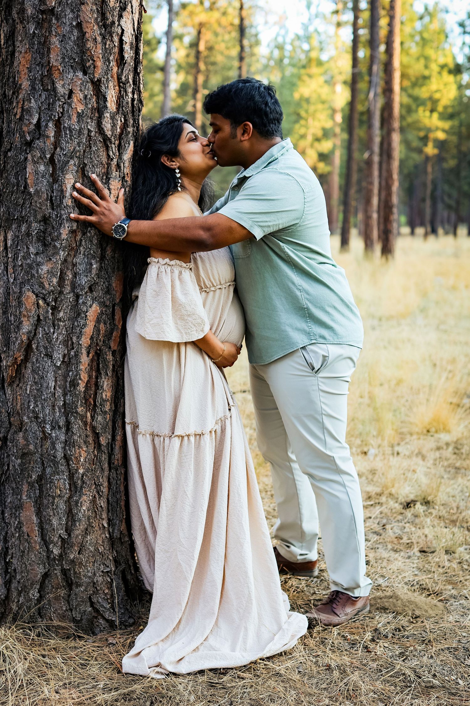 Pregnant couple sharing a kiss against a pine tree in a forest, woman in white maternity dress.