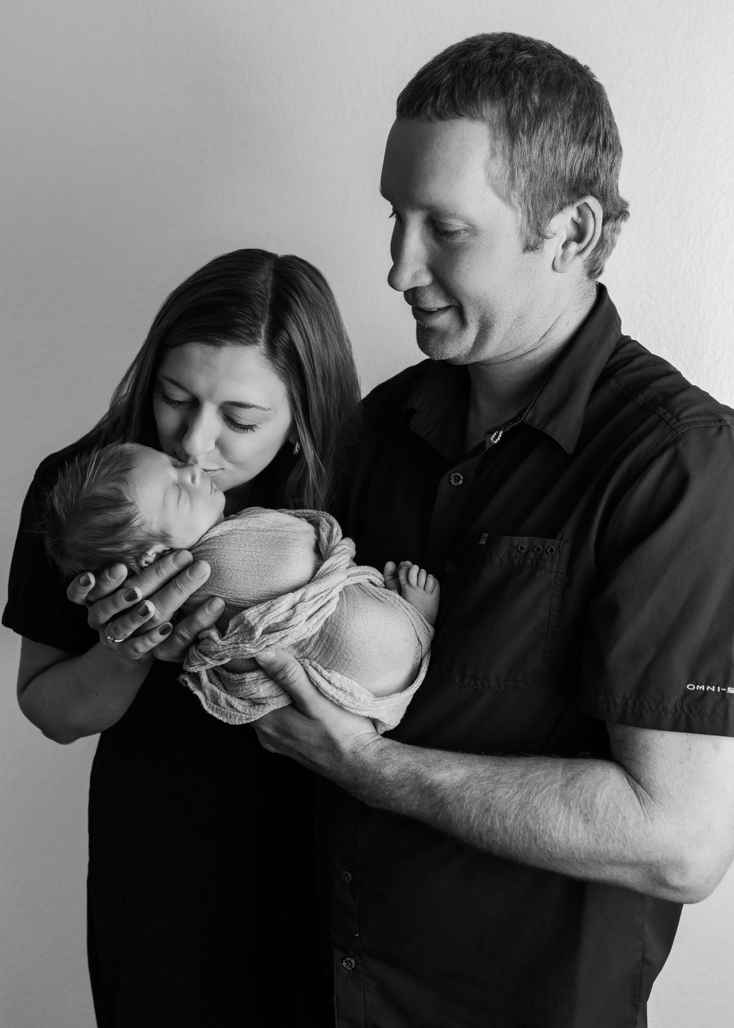 Black and white photo of new parents tenderly holding their swaddled newborn baby together.