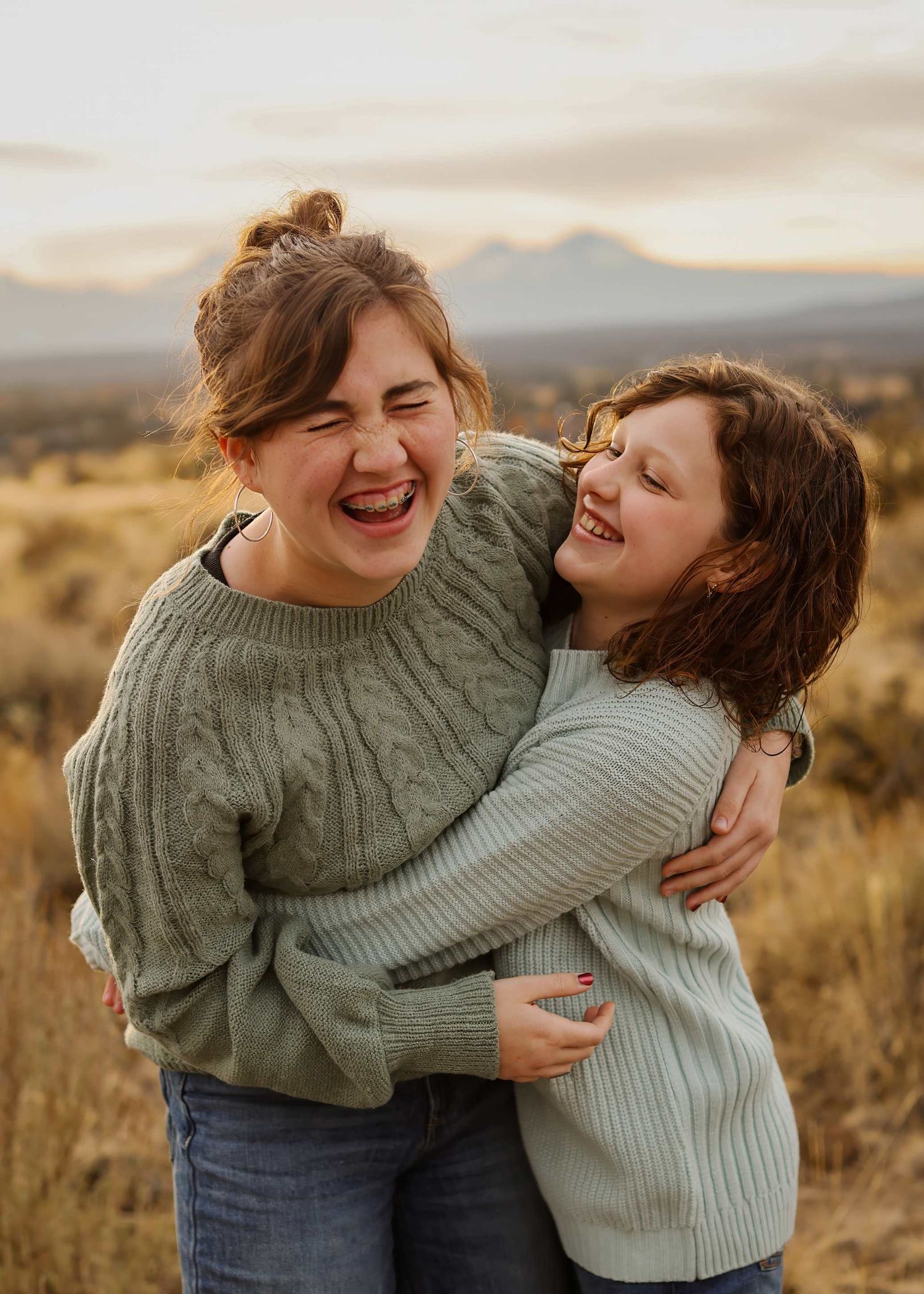 Two people in sweaters share playful embraces against a desert landscape at sunset.