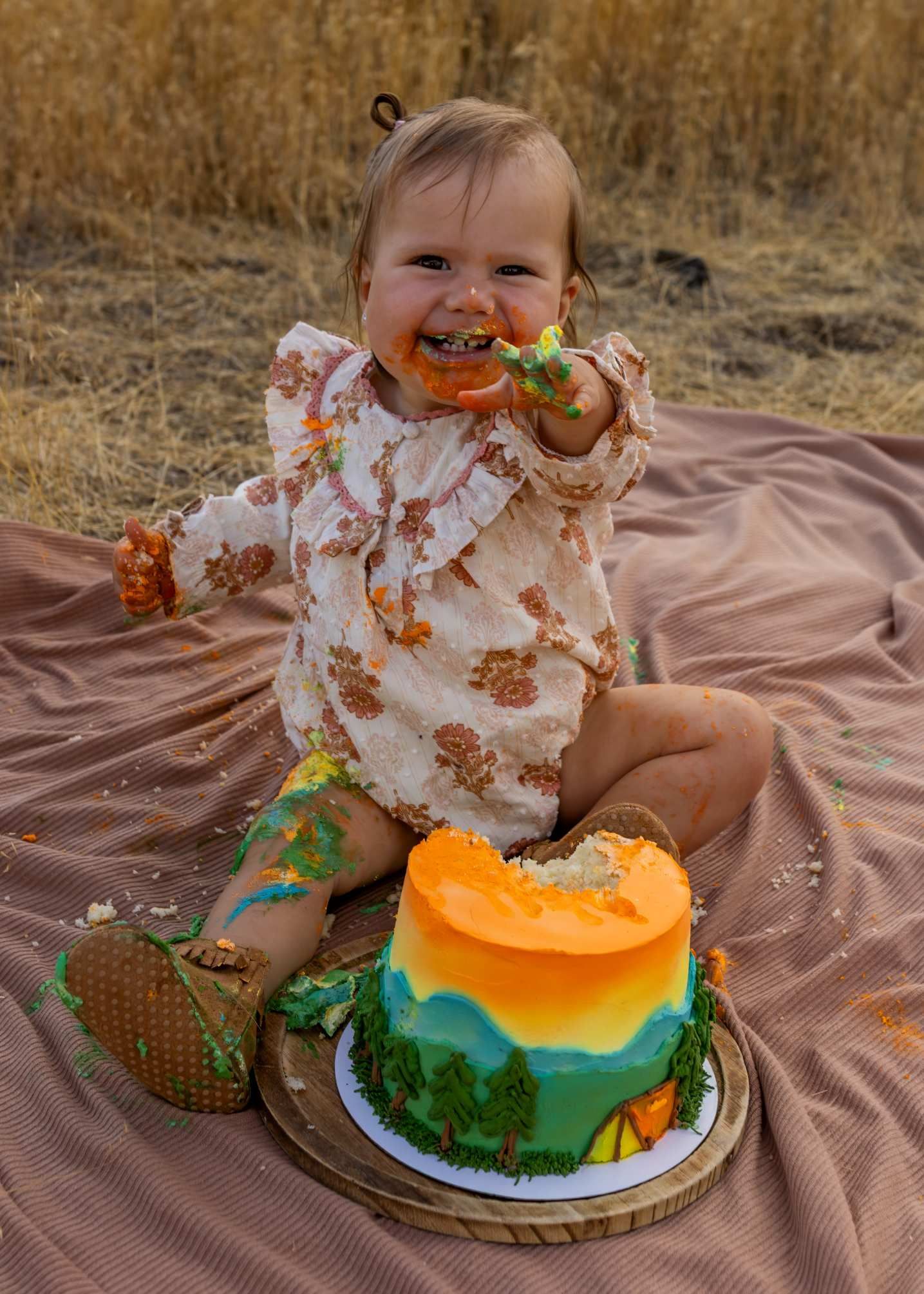 A messy cake smash photo session on a brown blanket featuring a decorated green and orange birthday cake.