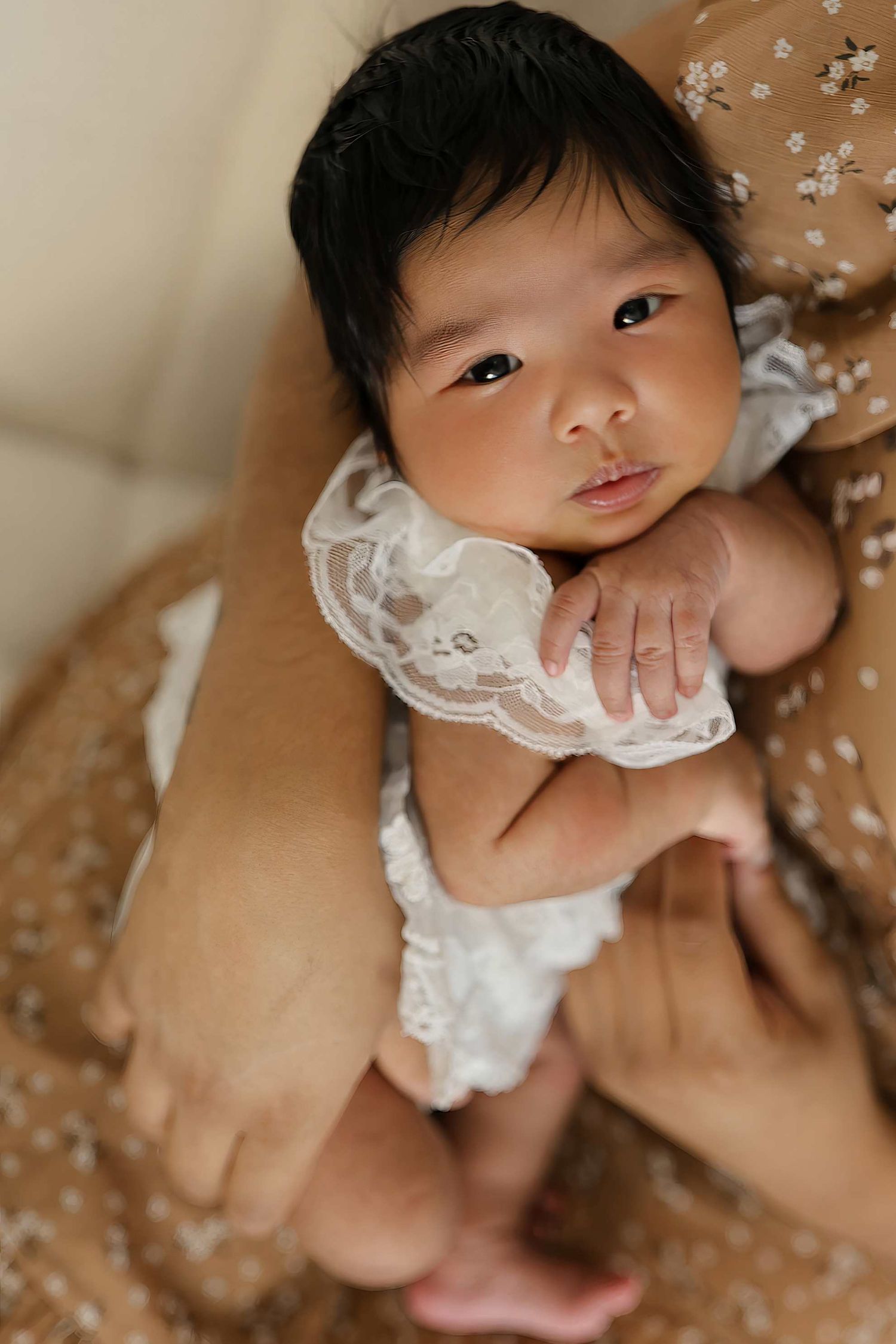 A baby wearing a delicate white lace dress lies on a beige surface with scattered flower petals around them. Redmond Oregon Studio