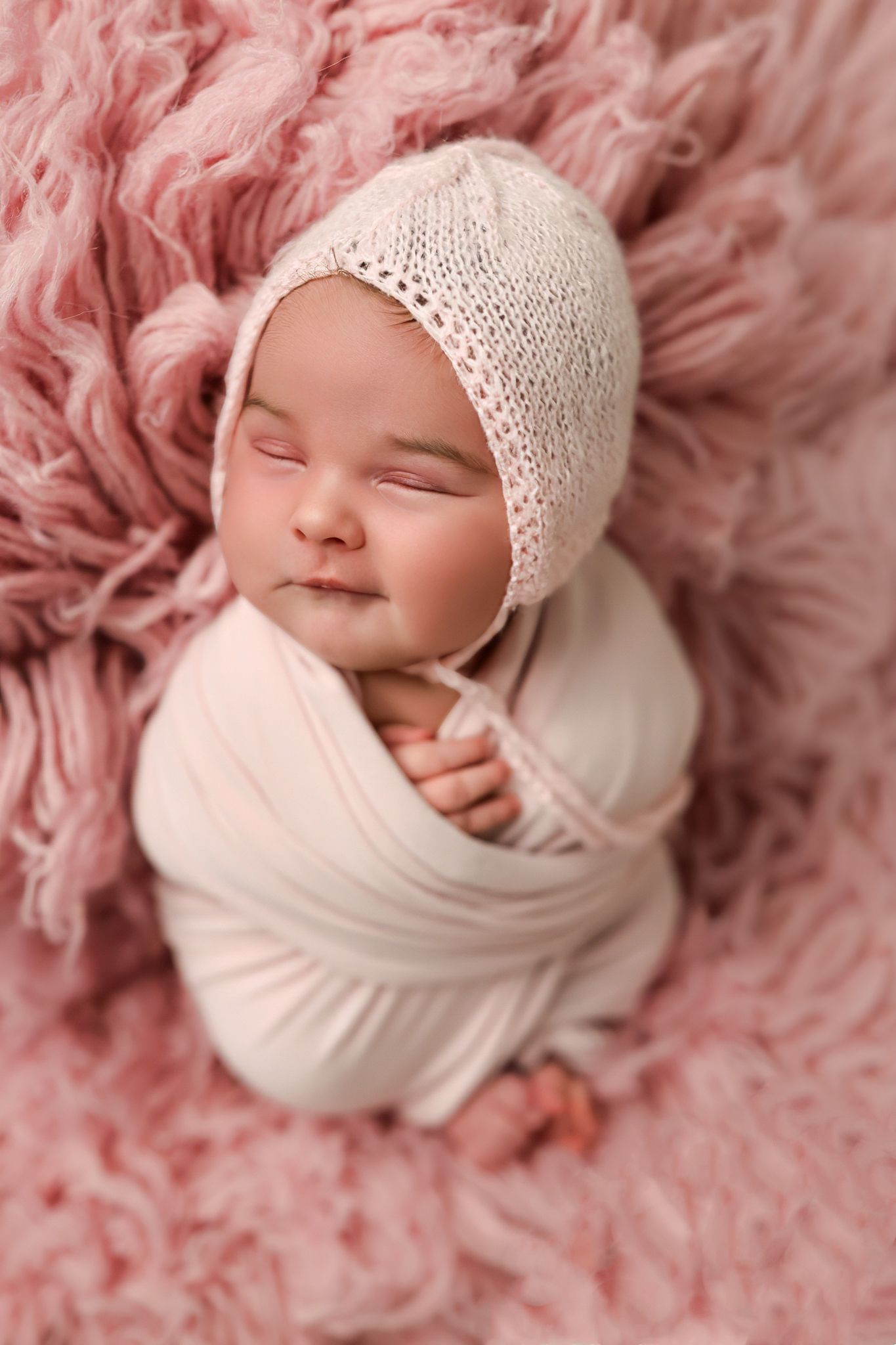 Sleeping baby wrapped in white swaddle with knit bonnet on pink fluffy backdrop.