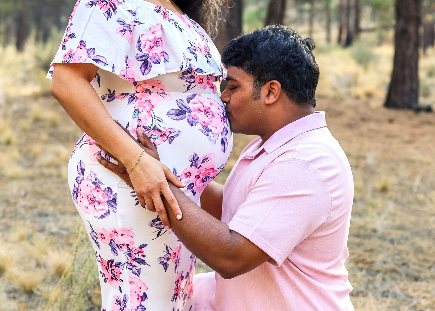 Man kneeling and kissing pregnant woman's belly outdoors during a maternity photo session.