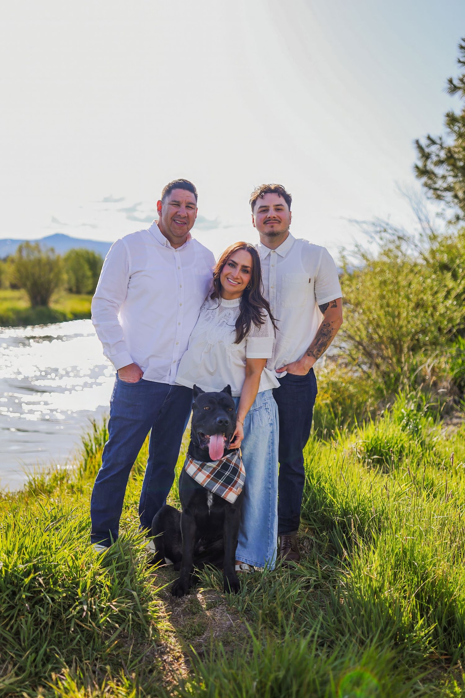 Family of two men and a woman pose outdoors by a river with their black dog wearing a bandana.