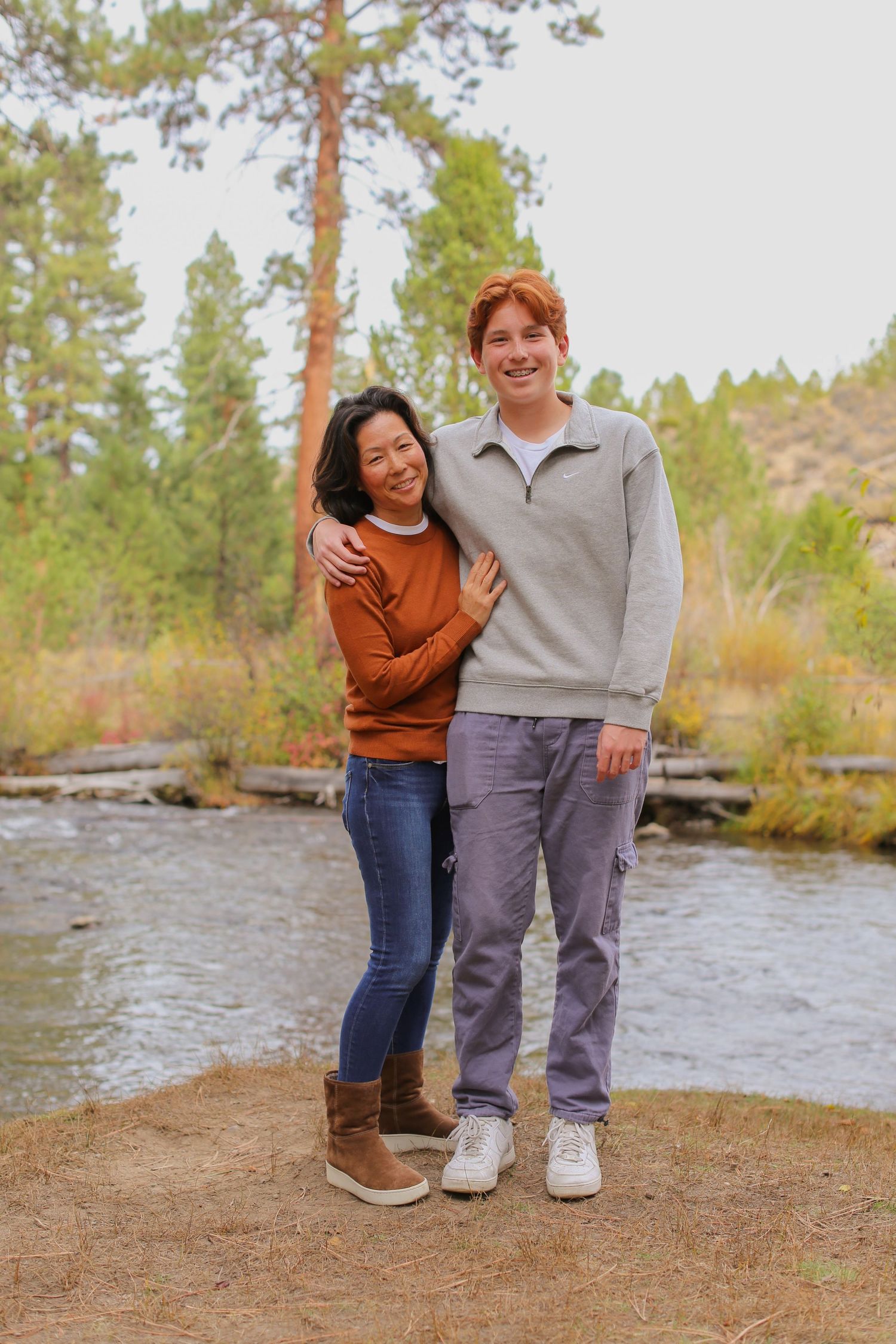 A woman and tall red-haired teen boy pose together near a river surrounded by pine trees and autumn foliage.