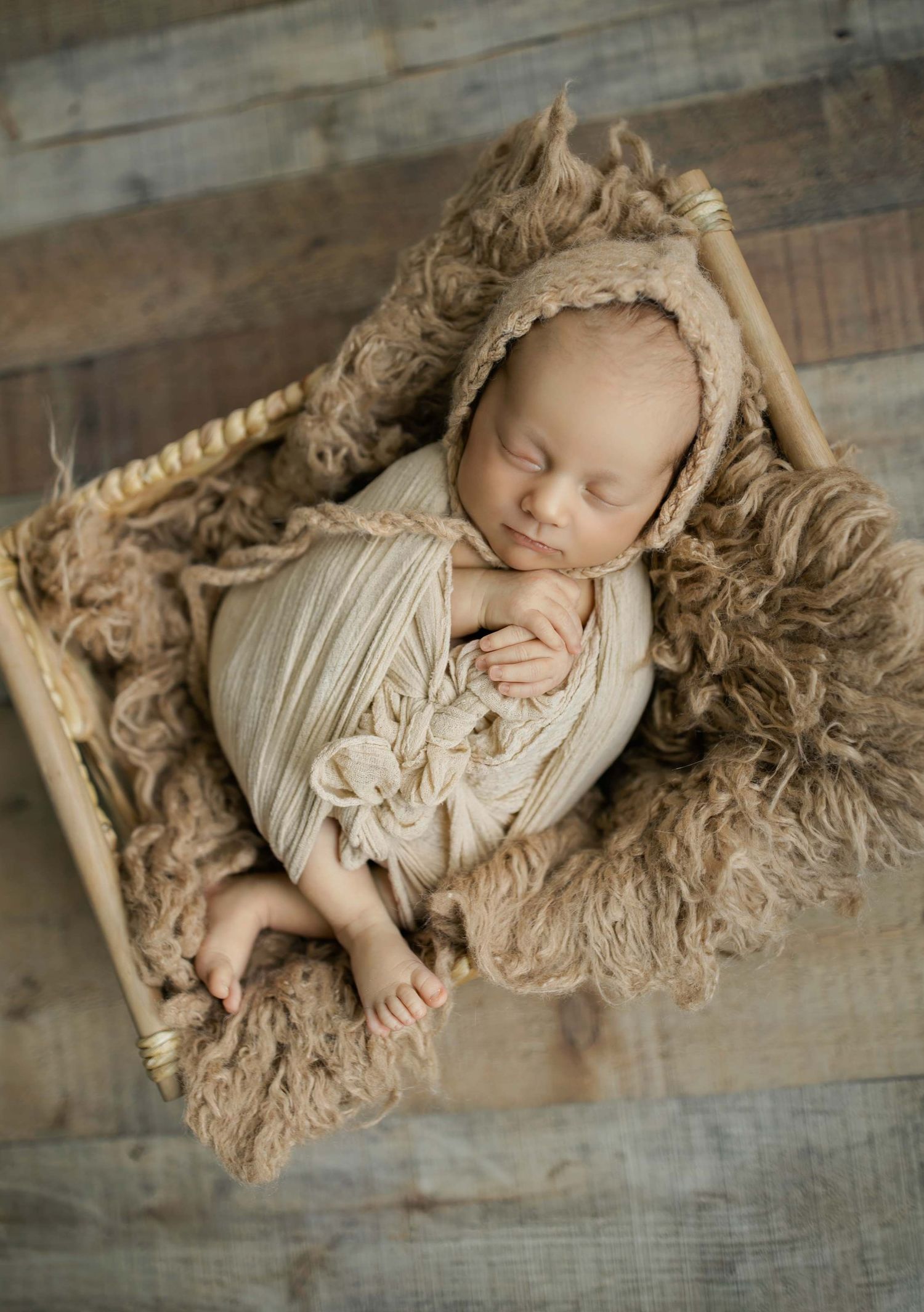 Sleeping newborn baby wrapped in cream swaddle with bonnet, nestled in rustic wooden crate with fluffy tan fur.