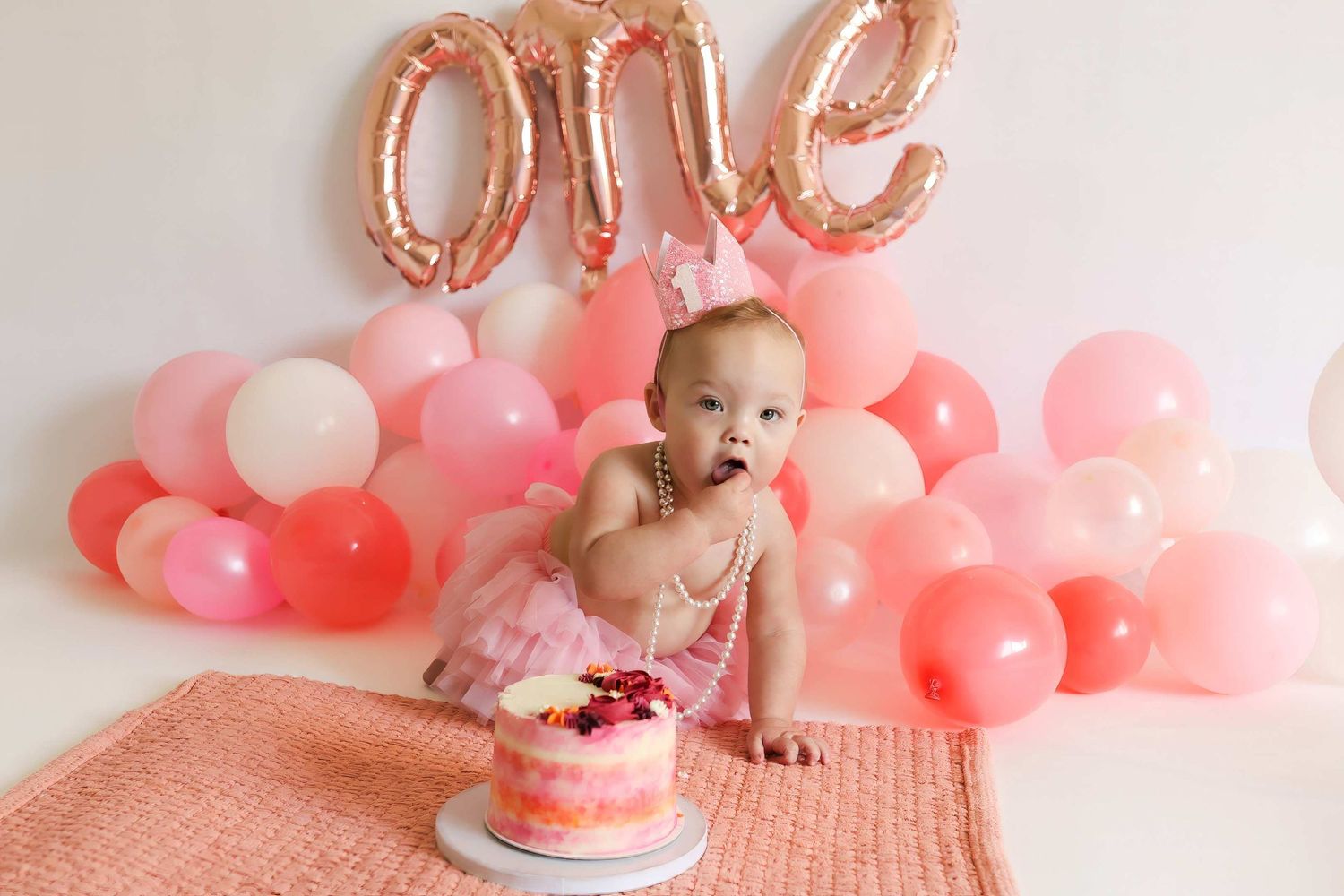 Baby girl celebrates first birthday smash cake with pink balloons and rose gold ONE balloon backdrop.