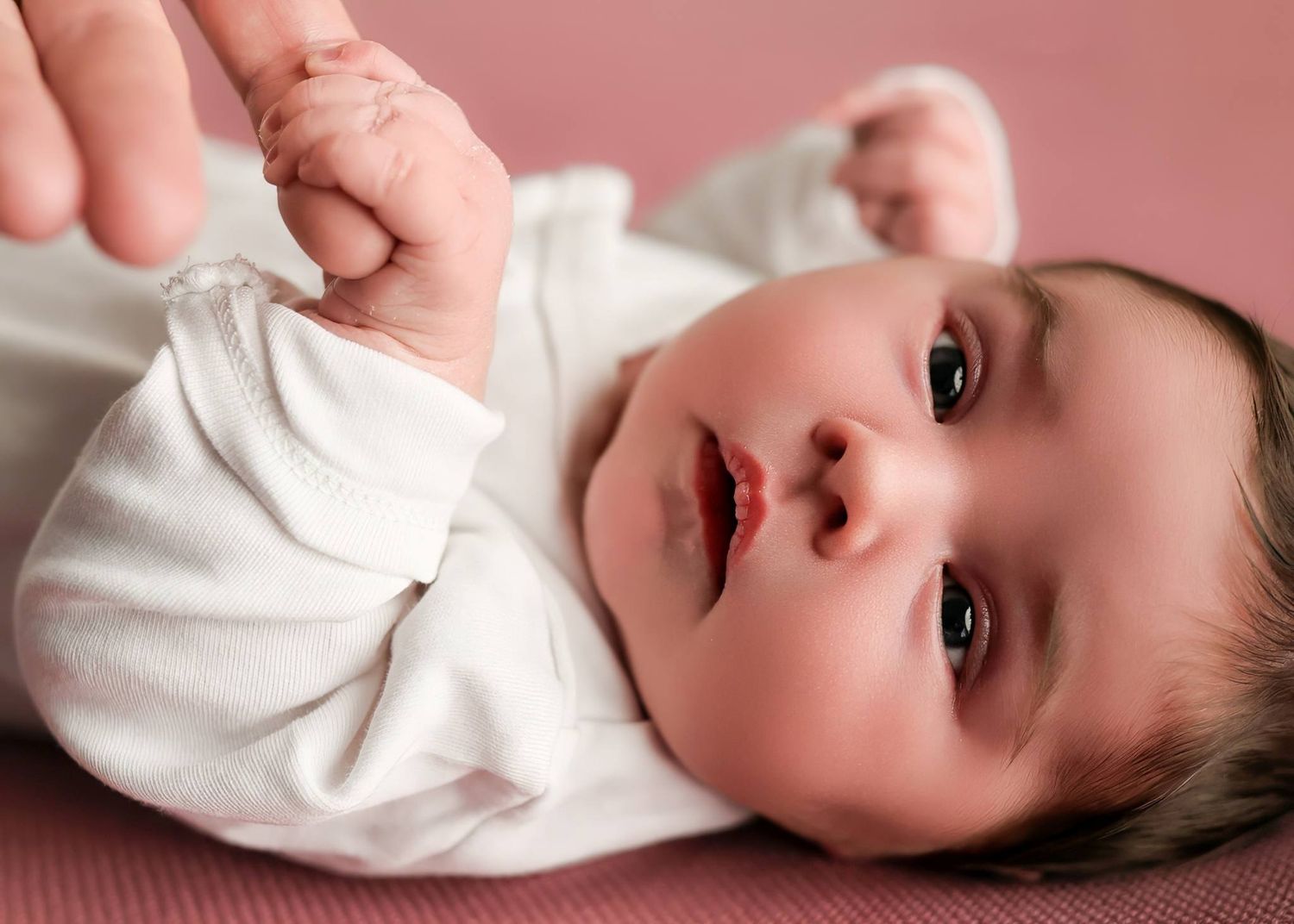 A baby in a white onesie lying on a pink blanket looking up at the camera.