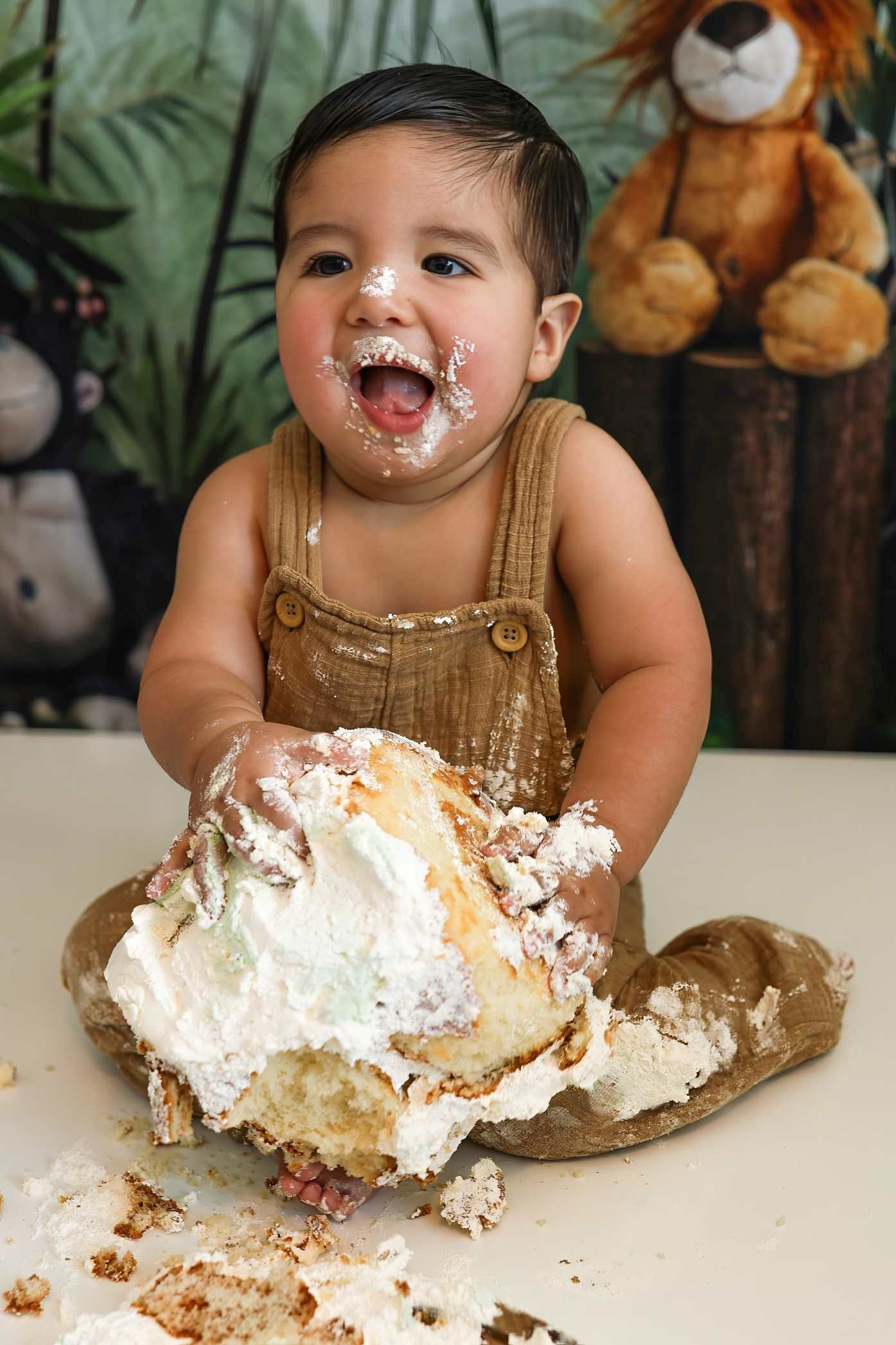 Baby in brown overalls enjoying a messy cake smash photoshoot with white frosting all over their face.