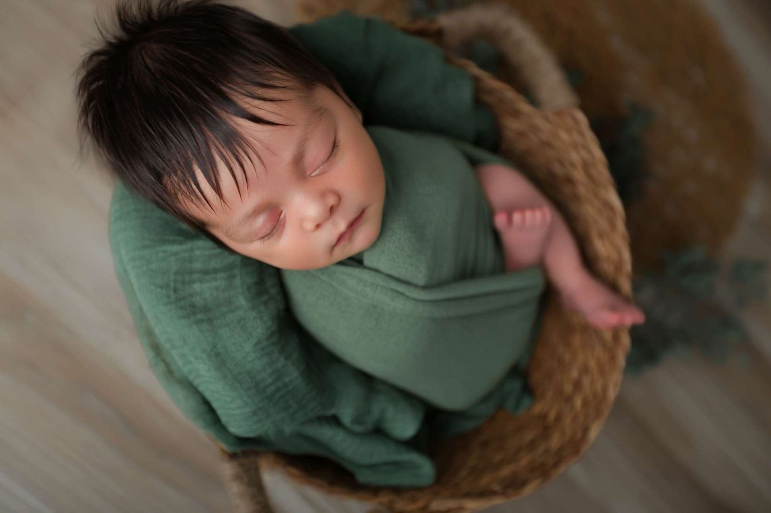 Newborn baby swaddled in green wrap sleeping peacefully in a woven basket.