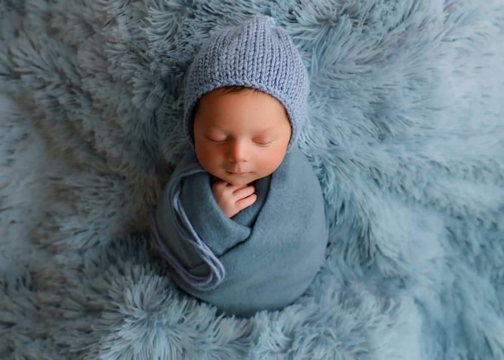 Newborn baby swaddled in blue wrap wearing knit bonnet, sleeping peacefully on soft blue fur blanket.