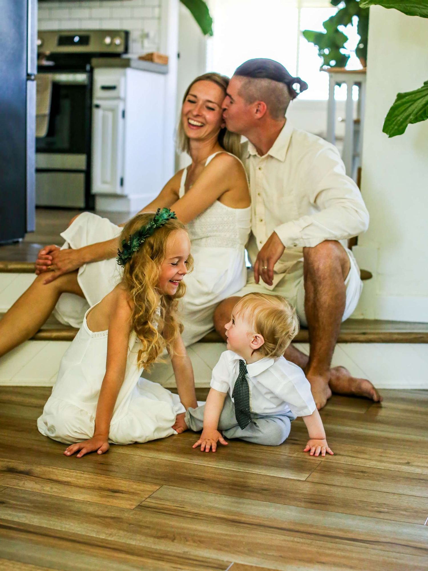 A joyful family of four in white outfits sits on indoor stairs as dad kisses mom while kids play.