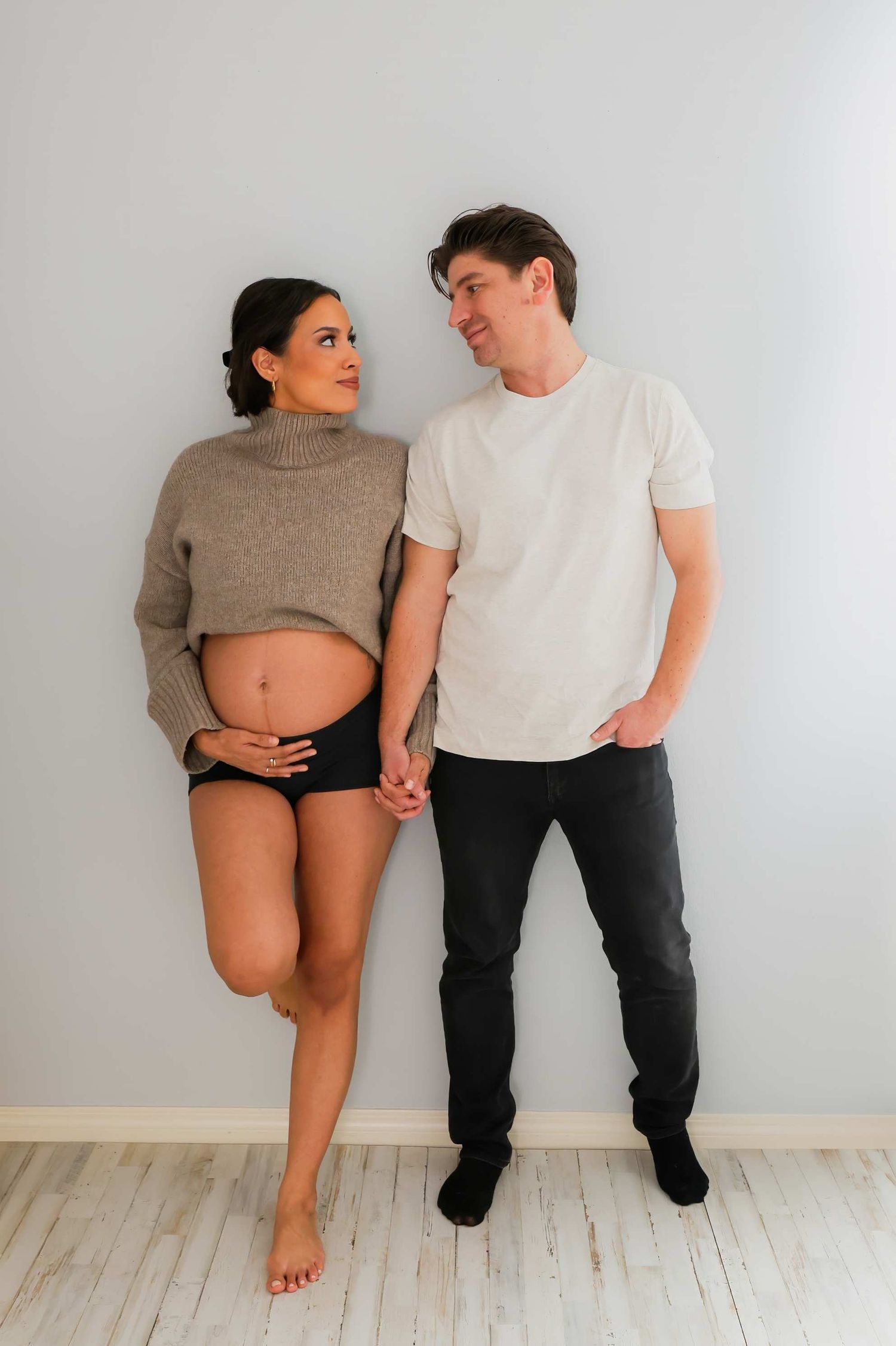 A couple poses against a white wall, with the woman in a cropped sweater showing her belly.