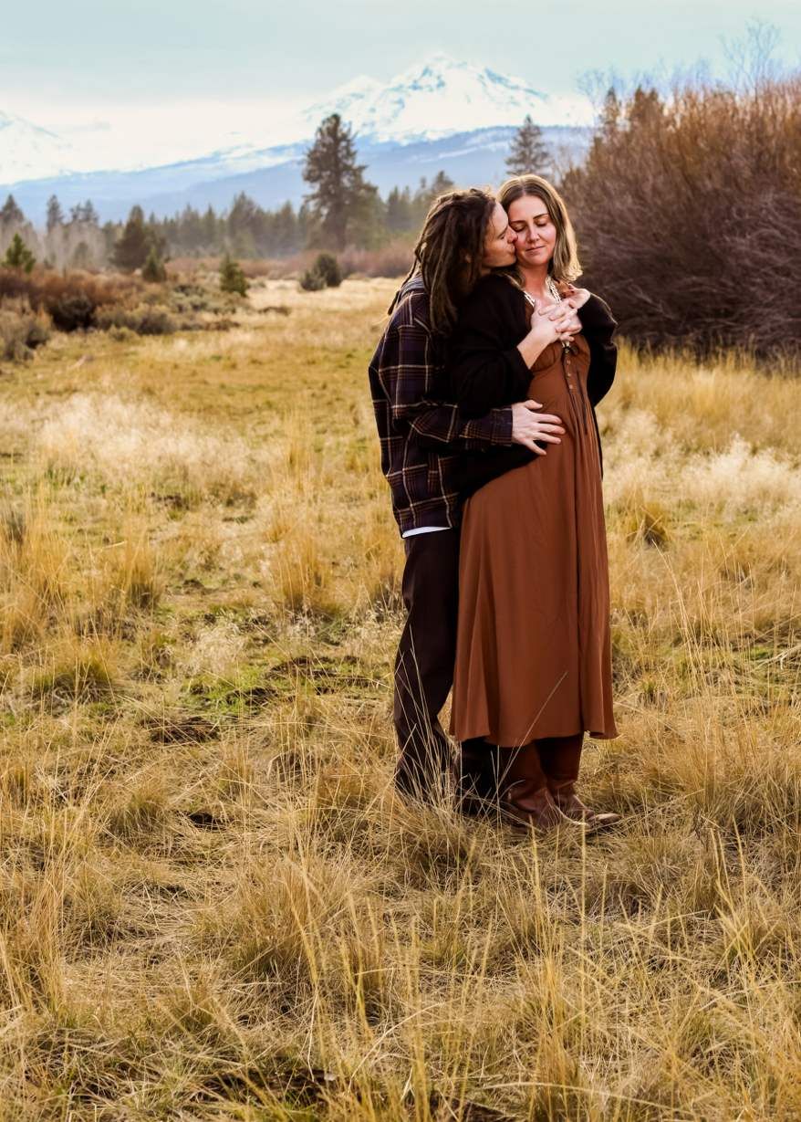Couple embracing in a rustic field with snow-capped mountains visible in the background.