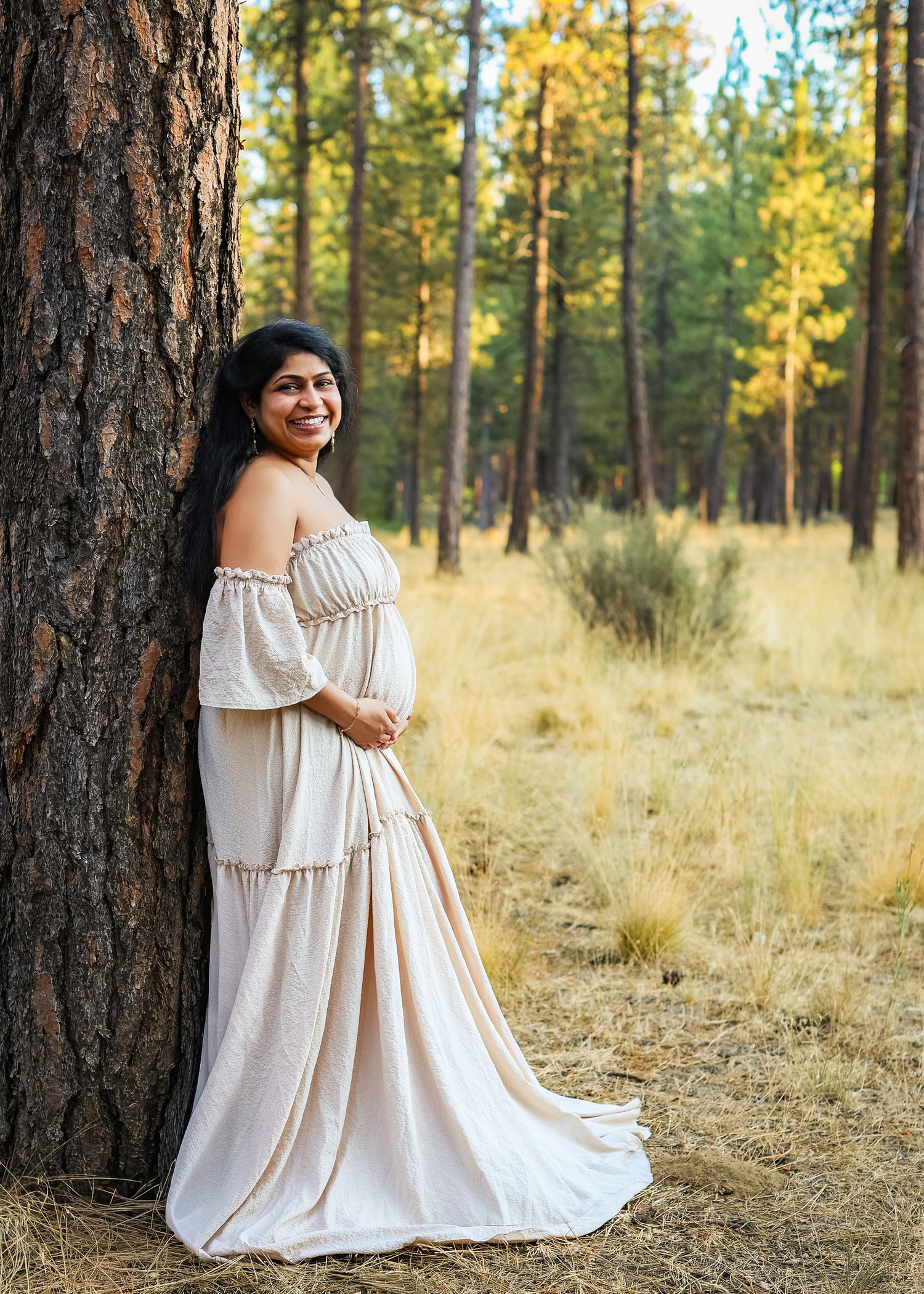 Smiling pregnant woman in white off-shoulder gown leaning against pine tree in golden forest meadow.