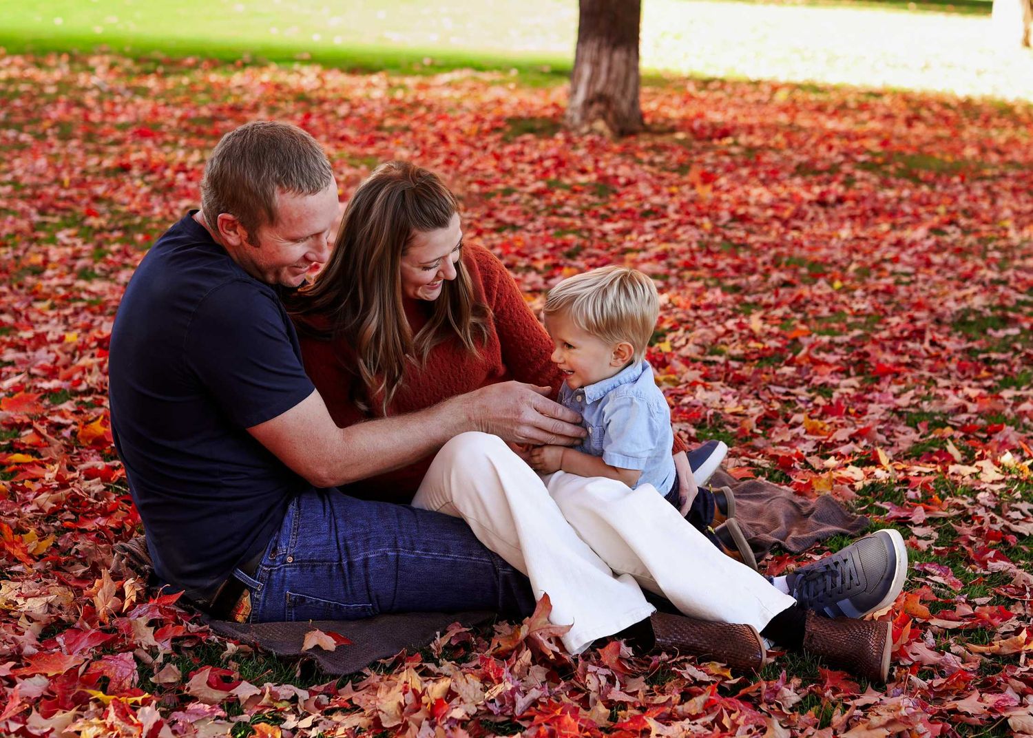 Family of three sitting in vibrant red autumn leaves in a park, parents smiling at toddler son.