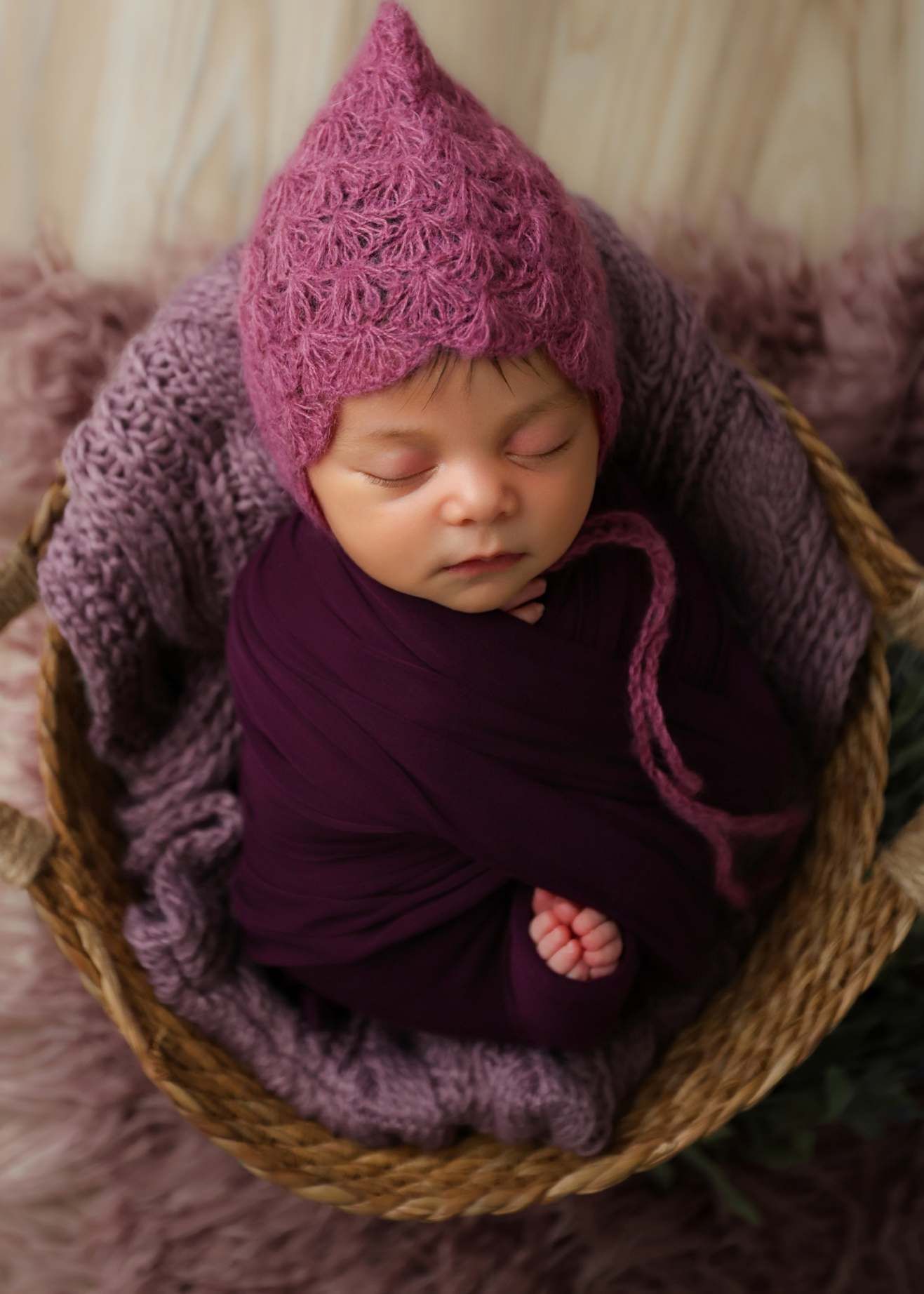 Sleeping newborn baby wrapped in deep purple swaddle wearing pink crochet pixie hat, nestled in wicker basket.
