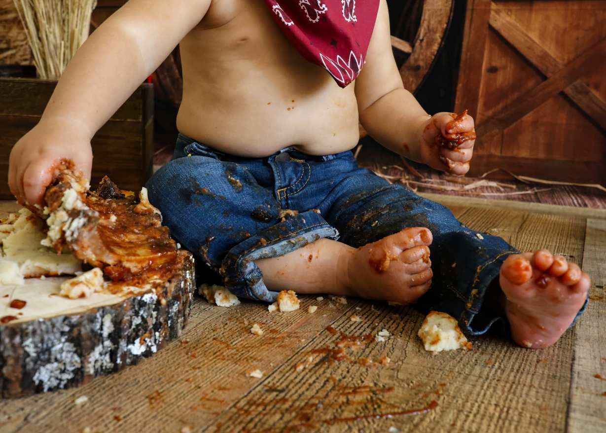 Messy toddler in jeans and bandana smashing cake on rustic wood floor during first birthday smash cake session.