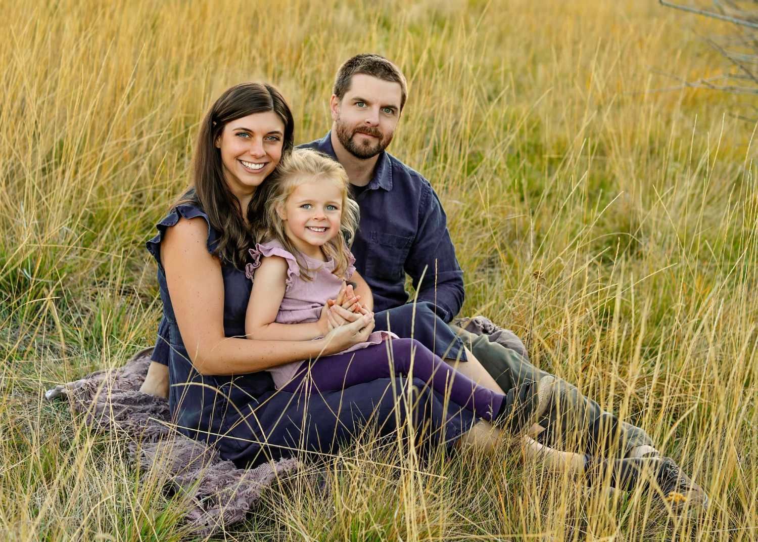 A smiling family of three sits in tall golden grass, parents flanking their young blonde daughter.