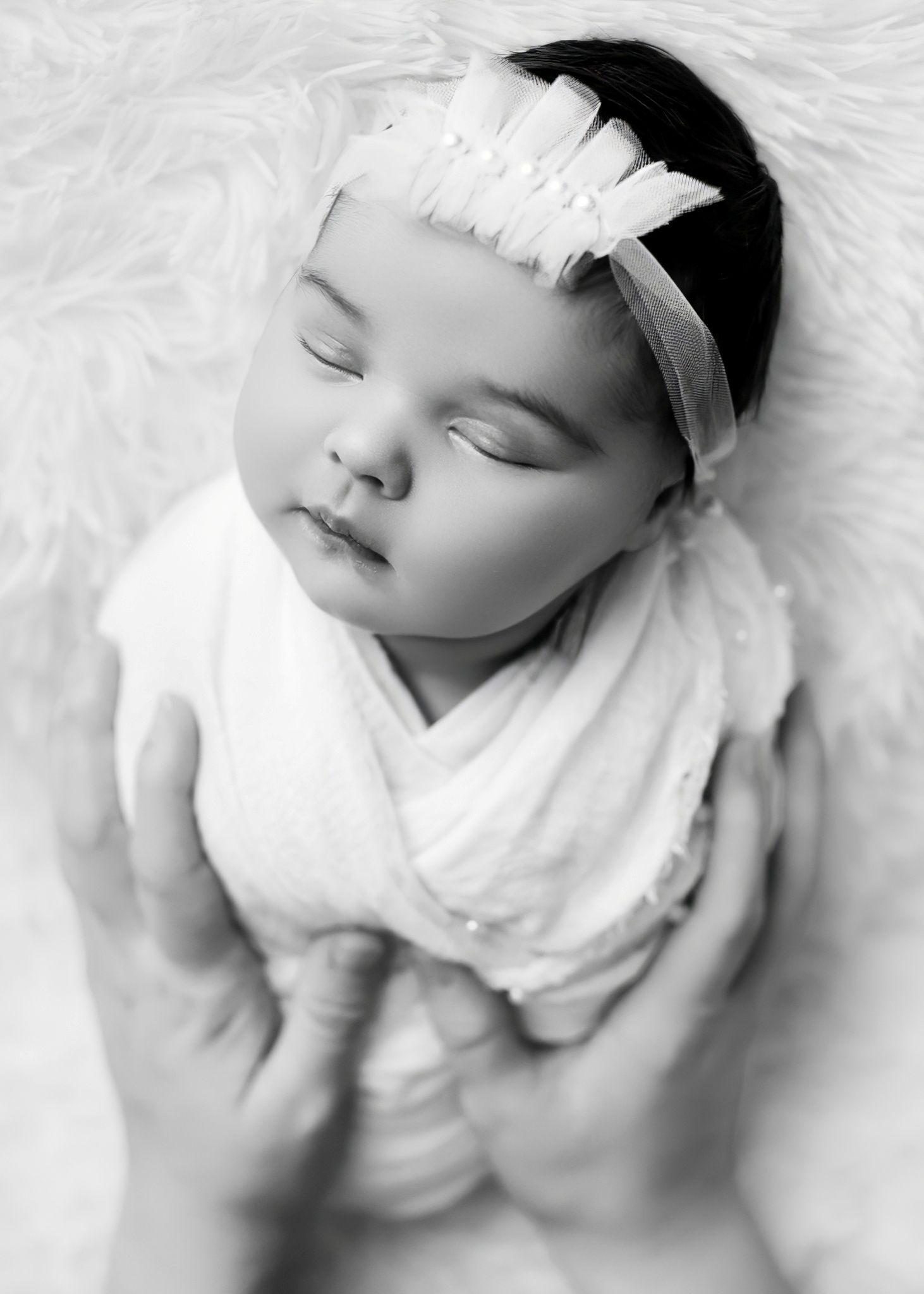 Black and white portrait of sleeping infant wearing white headband and wrap on fluffy background.