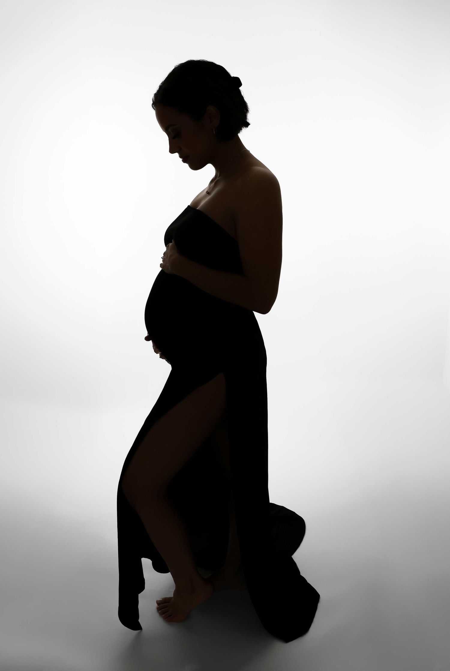 A silhouette of a pregnant woman in a flowing dress against a white background creates an artistic maternity portrait.