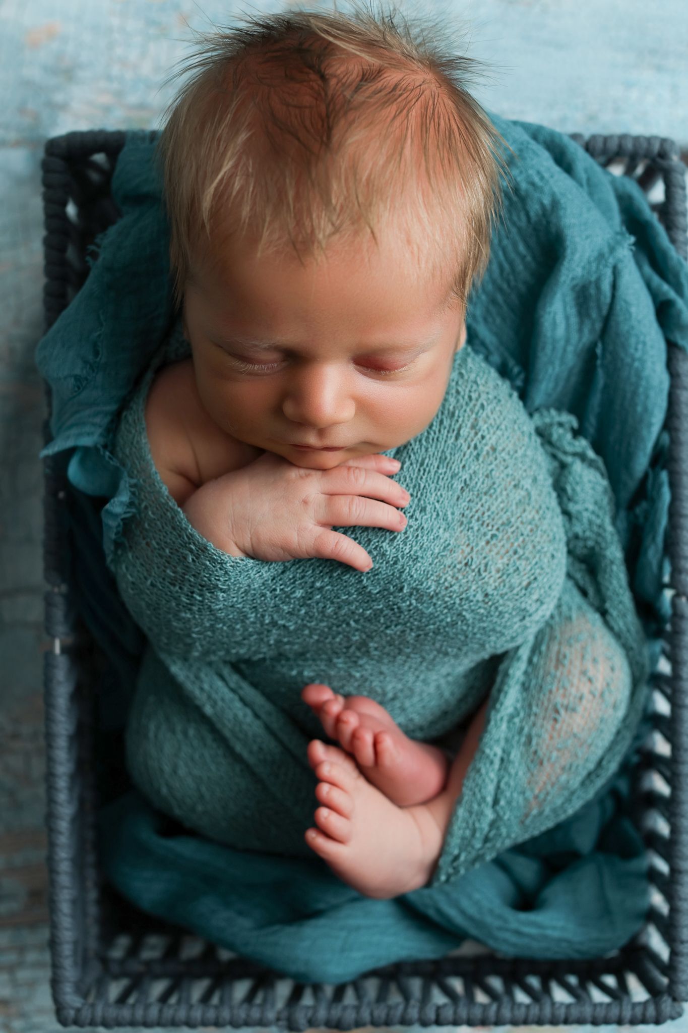 Newborn baby swaddled in teal wrap sleeping peacefully in a dark wicker basket on a blue background.