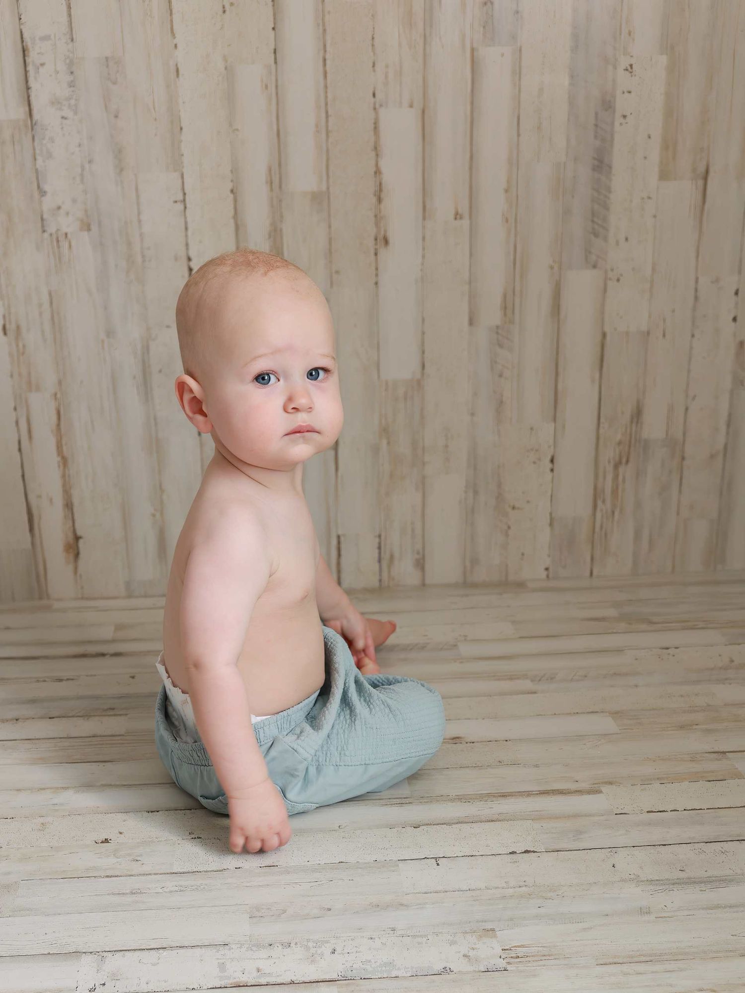 Baby wearing light blue pants sits on wooden floor against rustic white paneled wall in progressive movement shots.