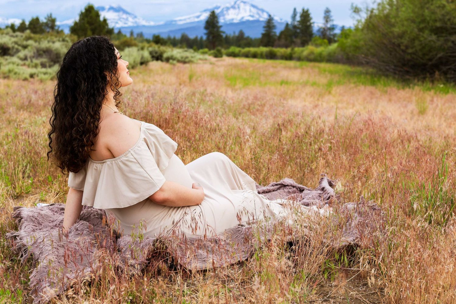 A person in a white dress rests in a field of tall grass with snow-capped mountains visible in the distance.