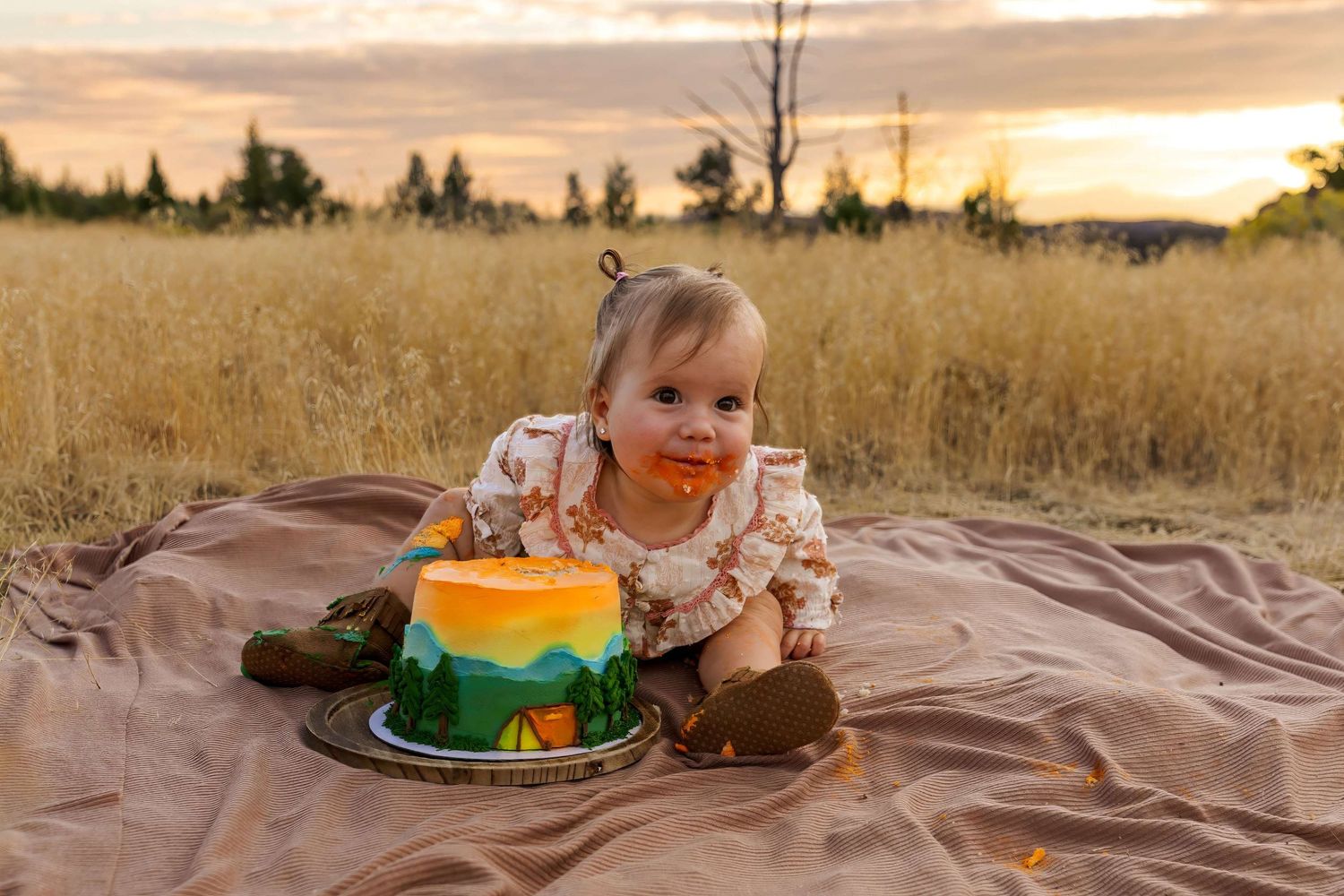 Baby's first birthday cake smash outdoors in golden field at sunset with colorful nature-themed cake.