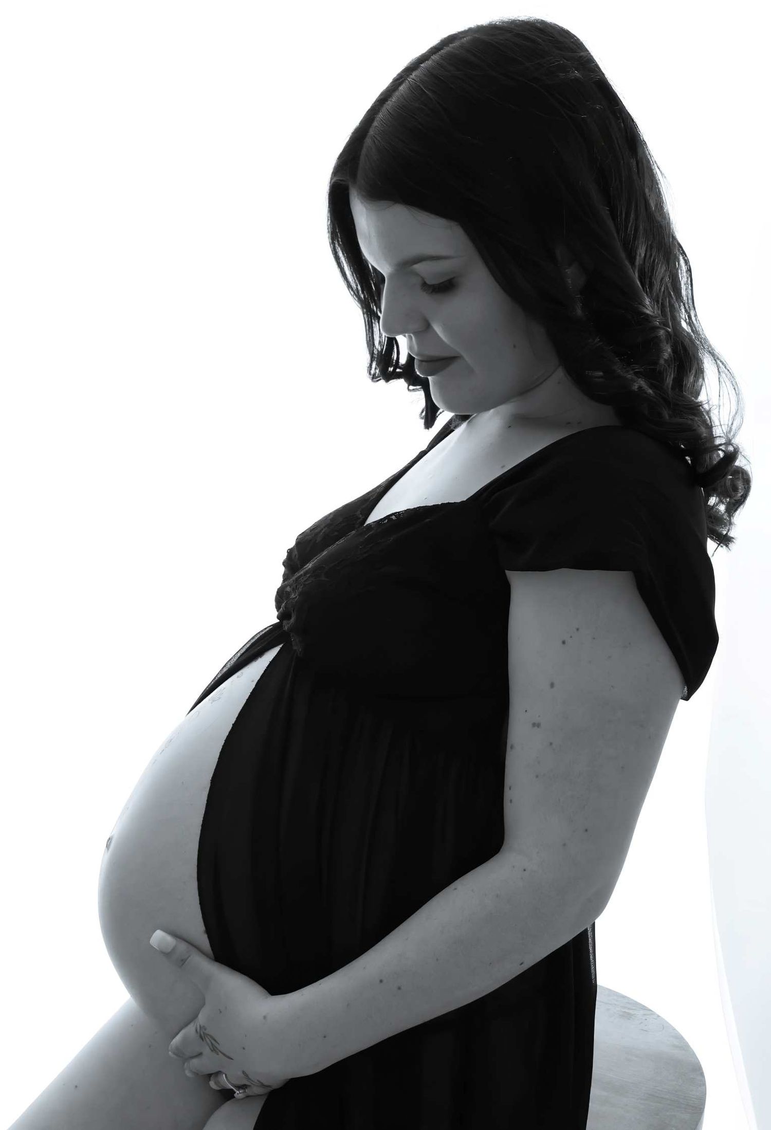 Black and white maternity photo of a pregnant woman in a dark dress cradling her baby bump while looking down.