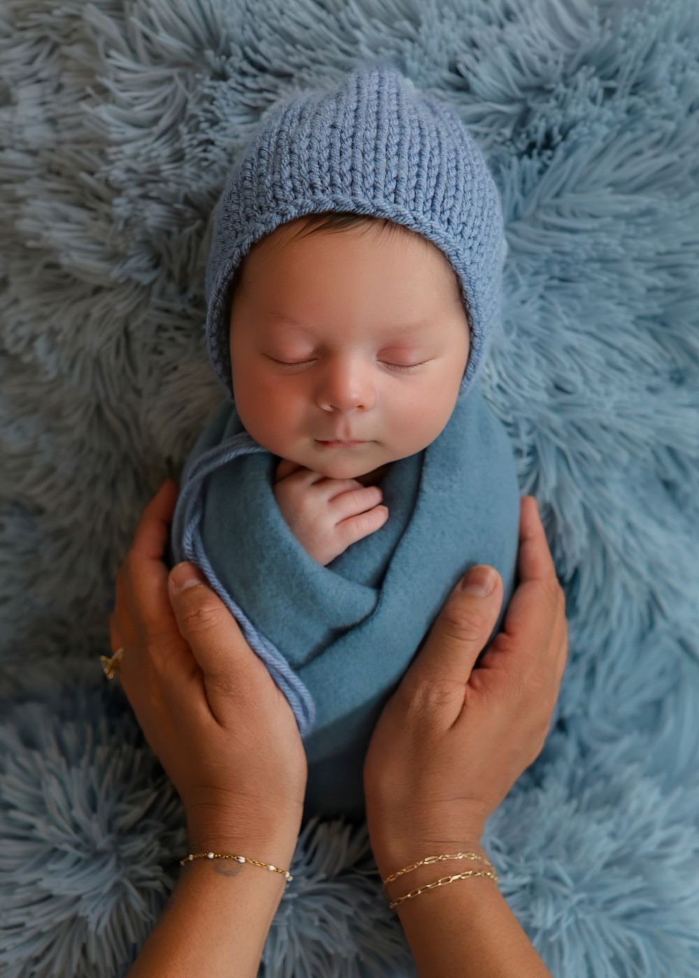 Sleeping newborn baby in blue knit bonnet and wrap cradled gently in adult hands on blue fluffy blanket.