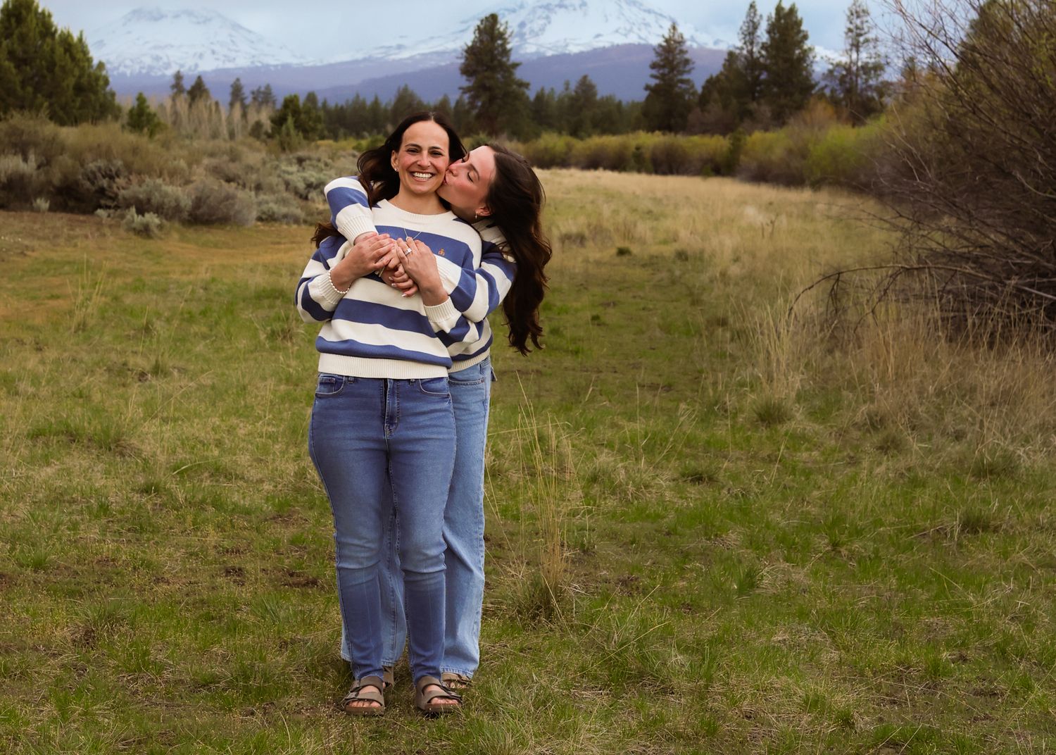 Two women hugging and smiling in a green meadow with mountains and trees in the background.