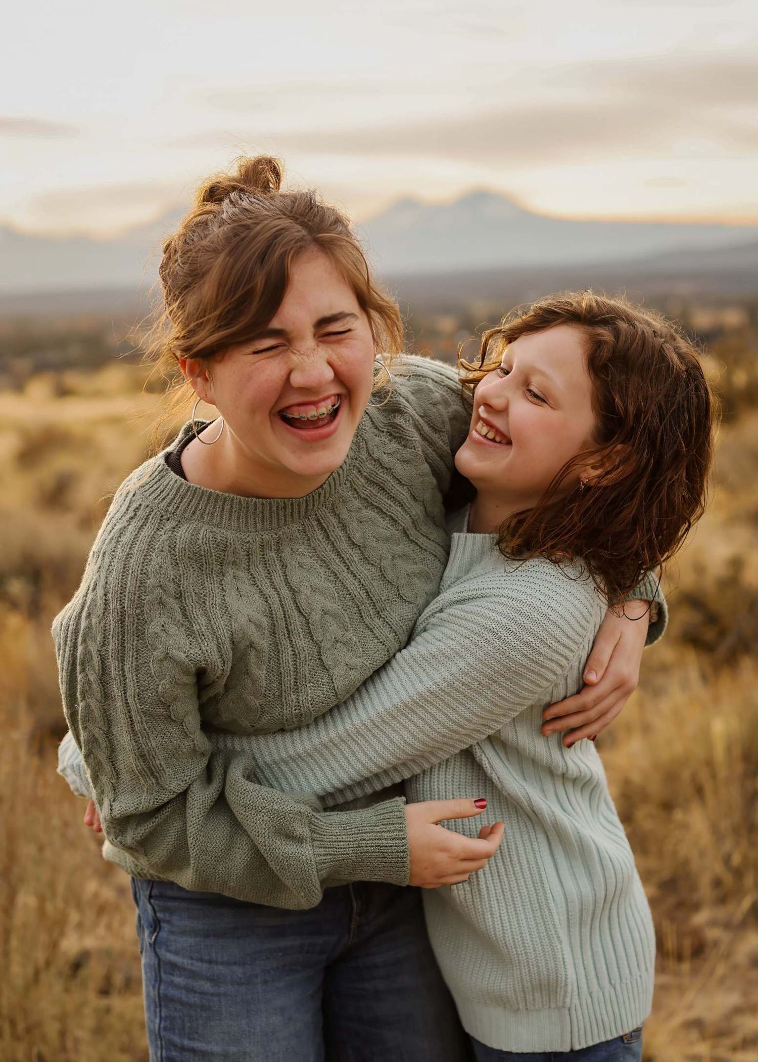 Two young girls in sage green sweaters laughing and hugging in a golden field with mountains at sunset. Redmond Oregon