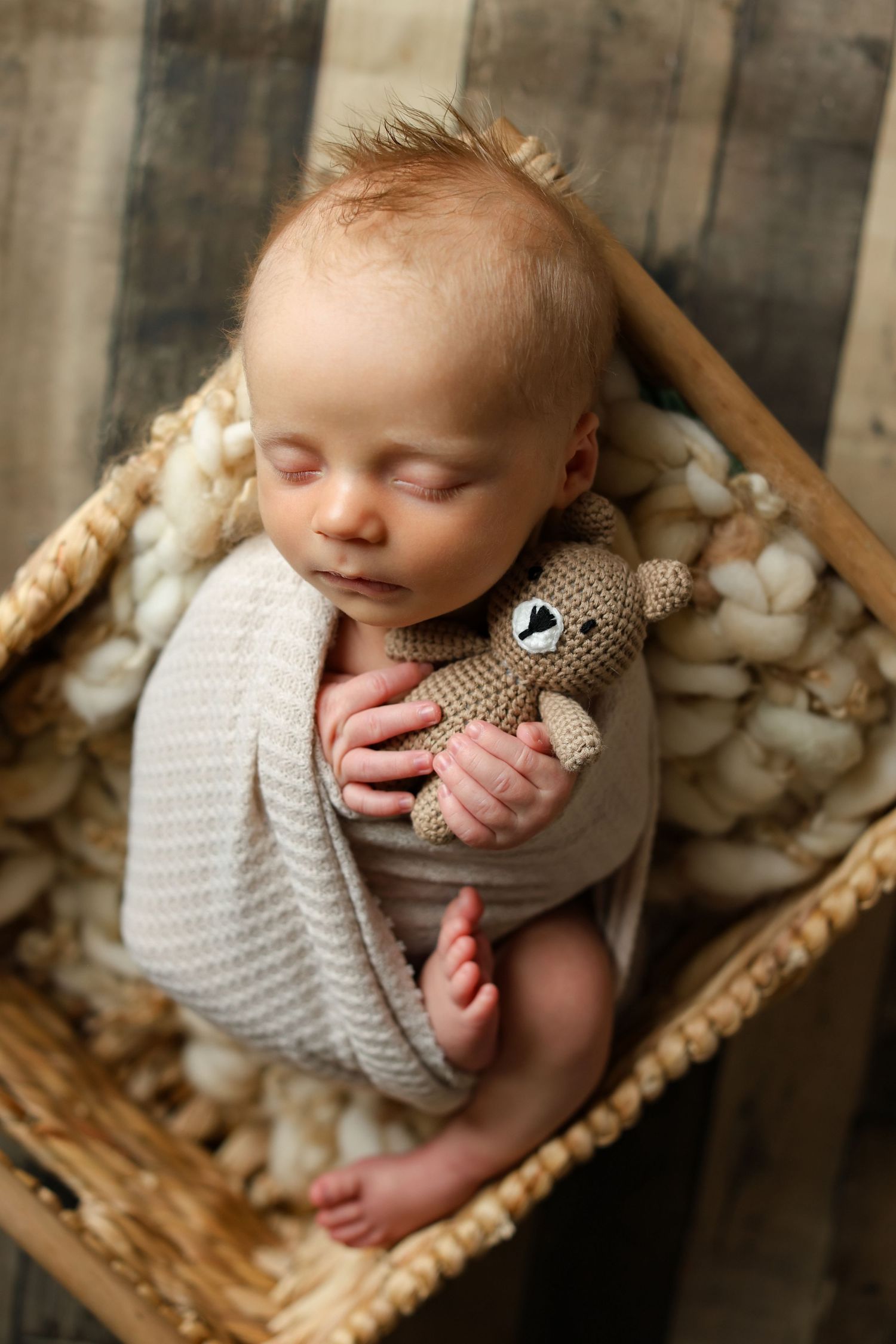 Sleeping newborn baby wrapped in a beige knit blanket, cradling a crocheted teddy bear inside a wicker basket.