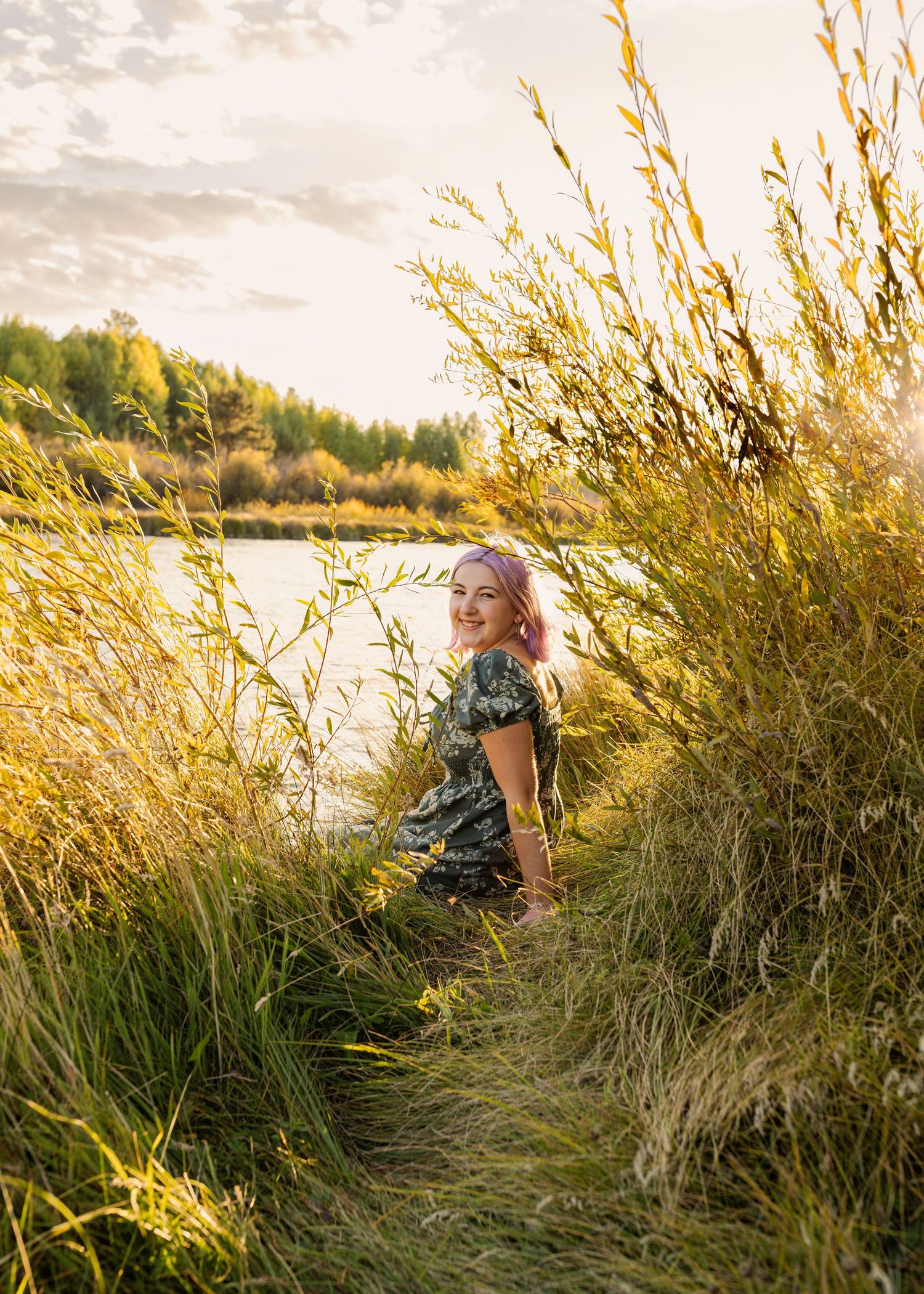 A smiling young woman with pink hair sits in tall golden grass near a sunlit lake at golden hour.
