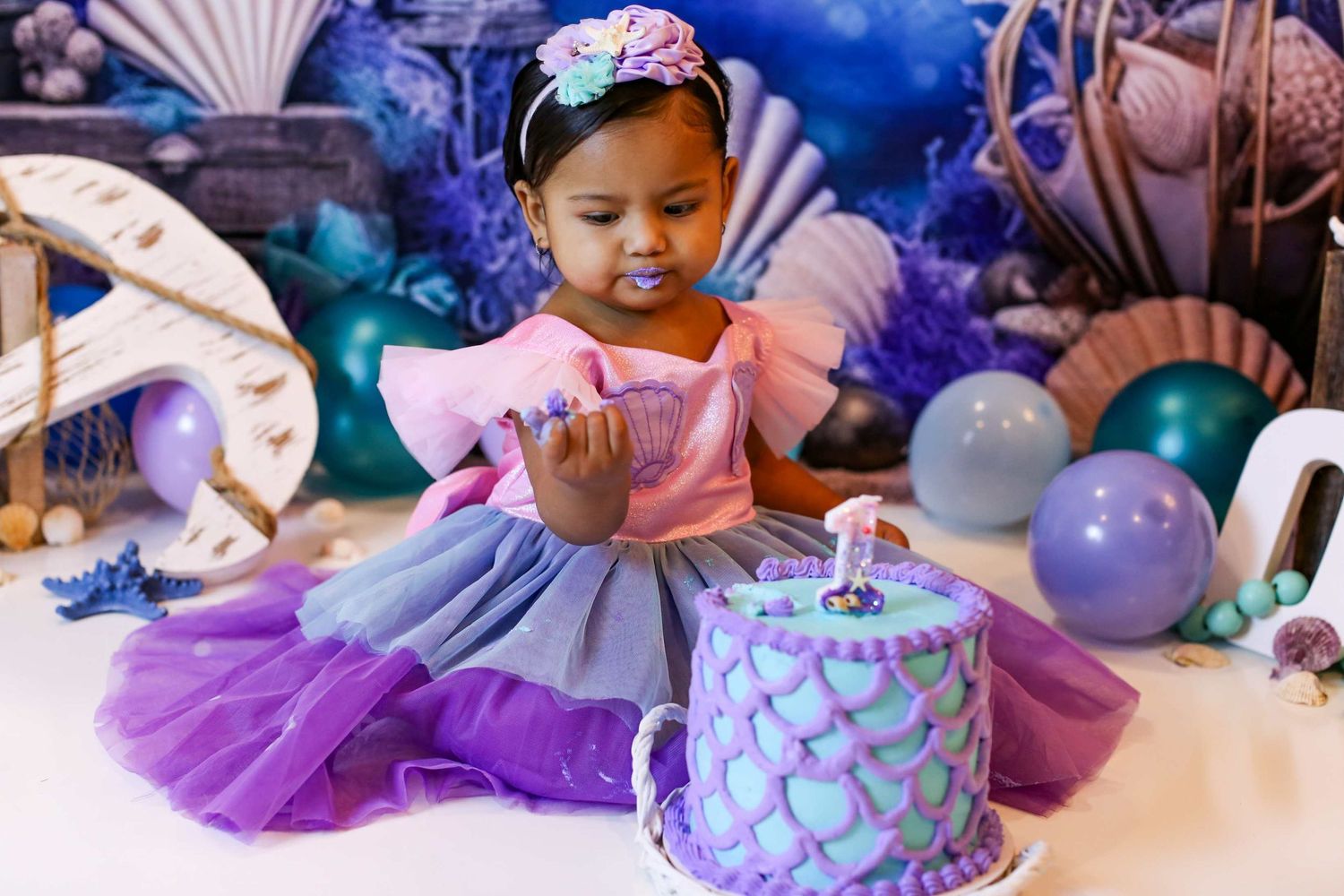 Toddler girl in purple tutu dress celebrating first birthday with mermaid themed cake and ocean decorations.